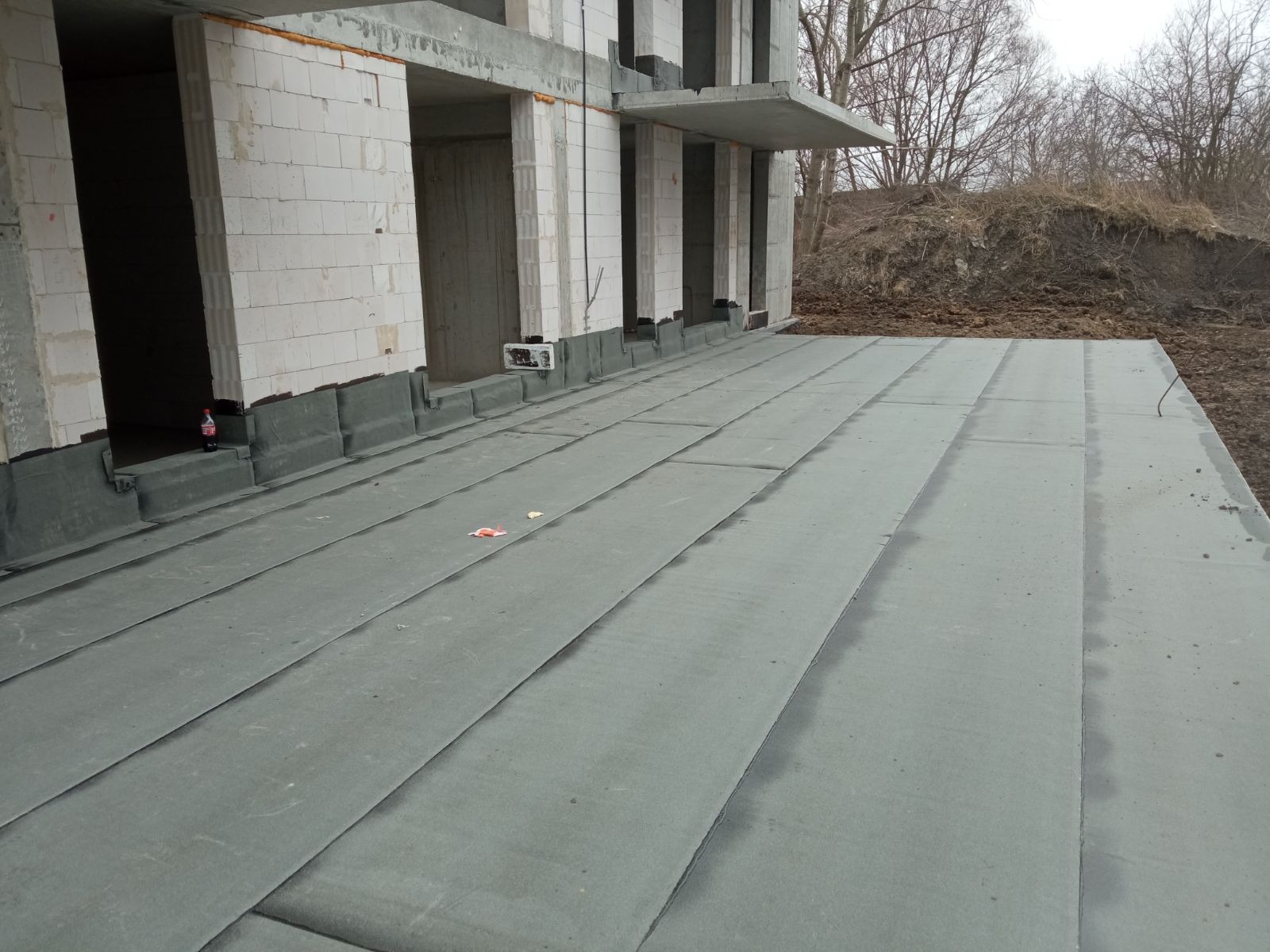Hydroizolacja płaskiego dachu budynku w stanie surowym otwartym, widoczne połączenia papy bitumicznej, w tle ściany z betonu komórkowego i częściowo zasypana ziemią skarpa.