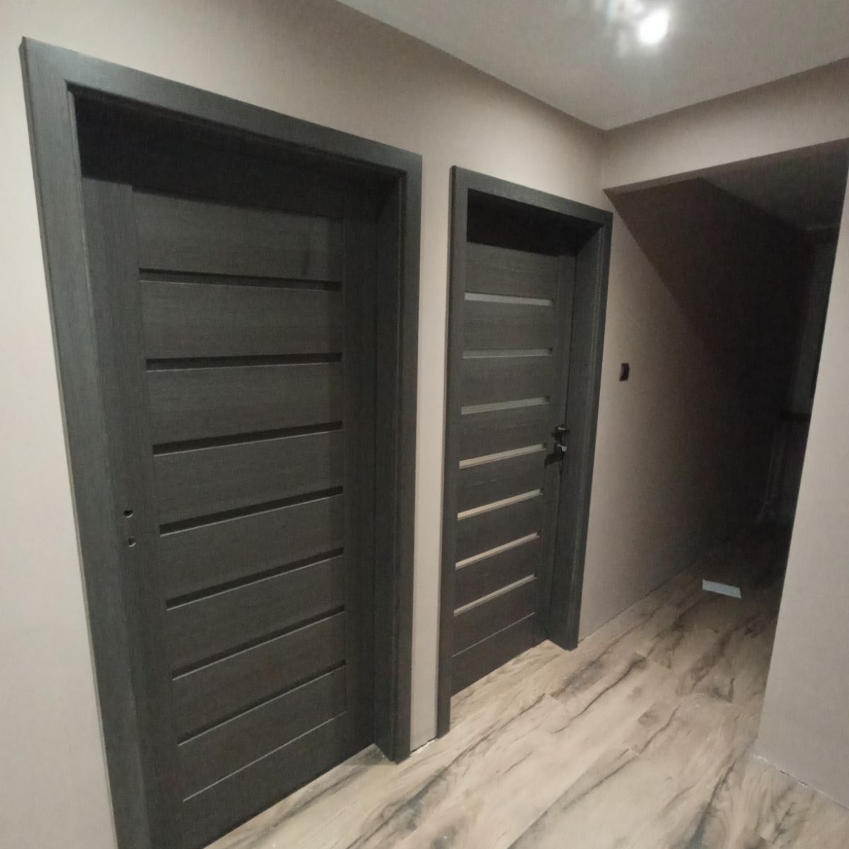 Dwa ciemnoszare, nowoczesne drzwi wewnętrzne z poziomymi panelami, zamontowane w korytarzu z podłogą imitującą marmur, widoczne otwory na klamki w jednych drzwiach.