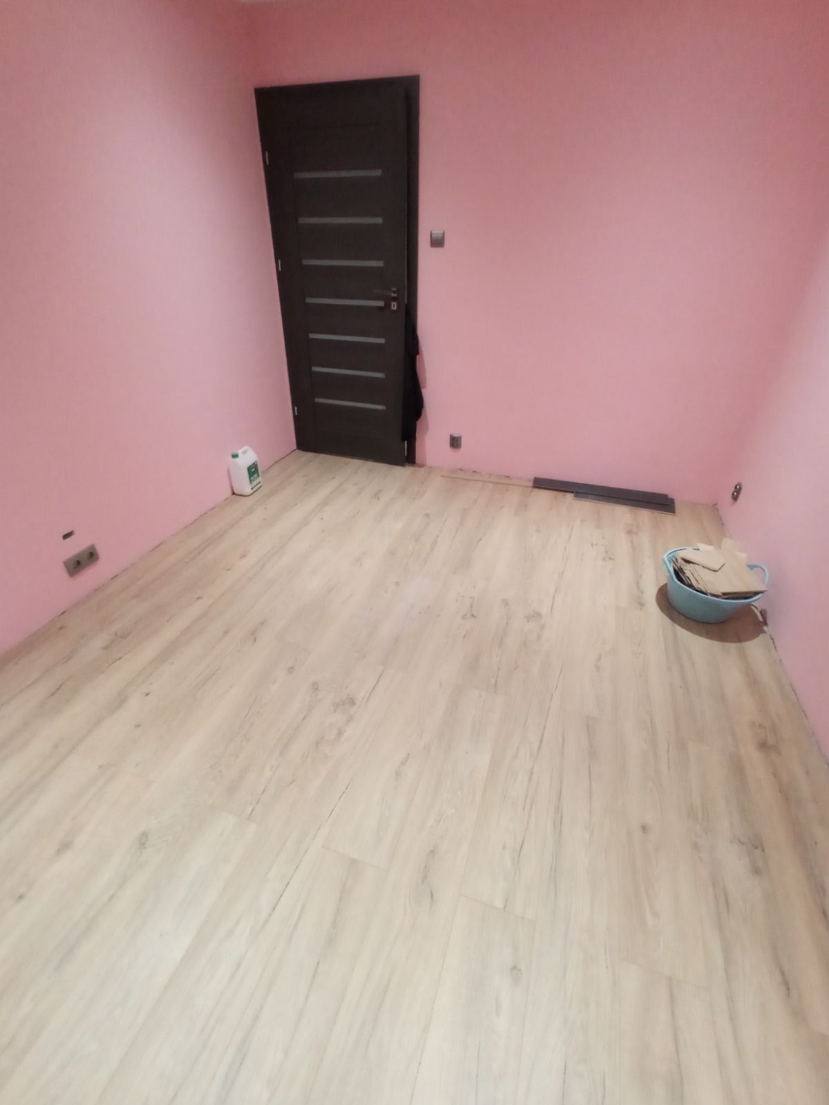 Pokój z nowo ułożonymi panelami podłogowymi o jasnym odcieniu drewna, różowe ściany, ciemne drzwi z poziomymi przeszkleniami, wiaderko z odpadami po montażu w rogu, fragmenty paneli przy ścianie.