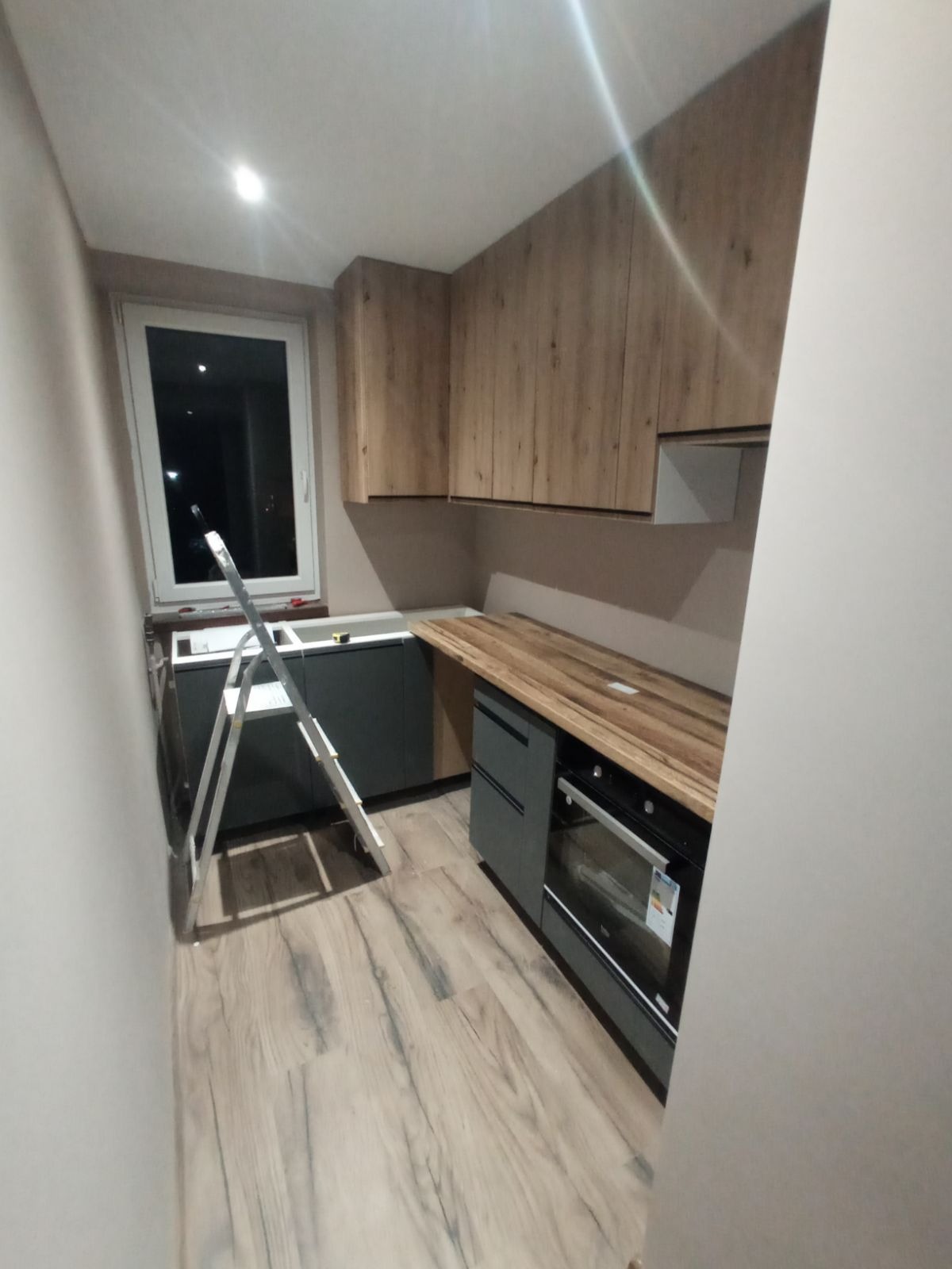 Montaż kuchni: widok na szarą zabudowę dolną z piekarnikiem, drewniany blat i szafki wiszące z imitacją drewna, drabina oparta o okno.