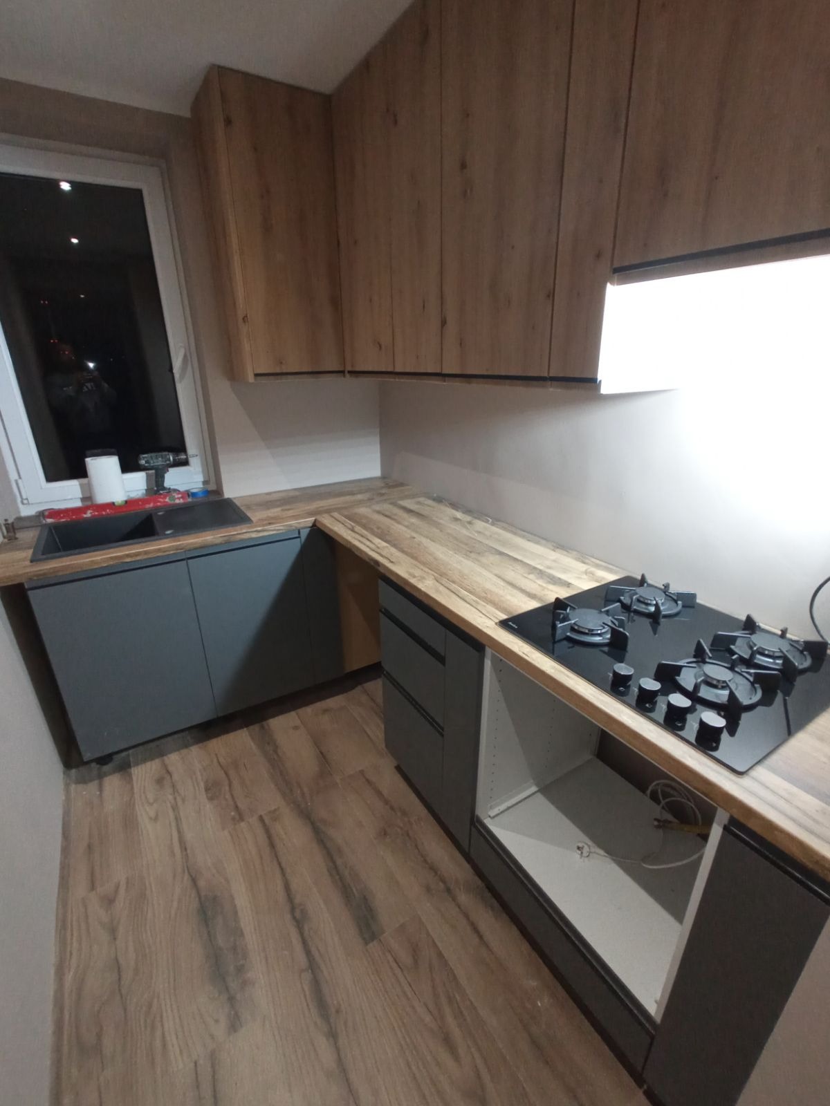 Nowoczesna kuchnia z drewnianymi szafkami wiszącymi, szarymi szafkami dolnymi, blatem imitującym drewno, czarną płytą gazową i zlewozmywakiem w rogu, widoczne narzędzia na parapecie okiennym.