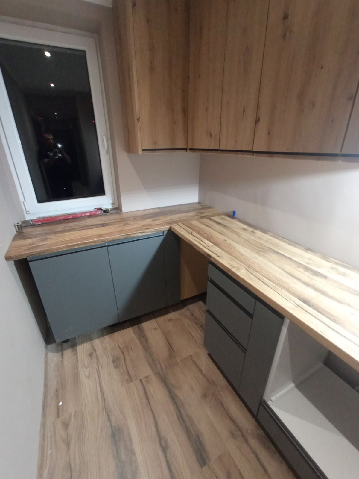 Nowoczesna kuchnia z szarymi szafkami dolnymi i drewnianymi górnymi, drewniany blat roboczy, widok z boku, podłoga z paneli imitujących drewno.