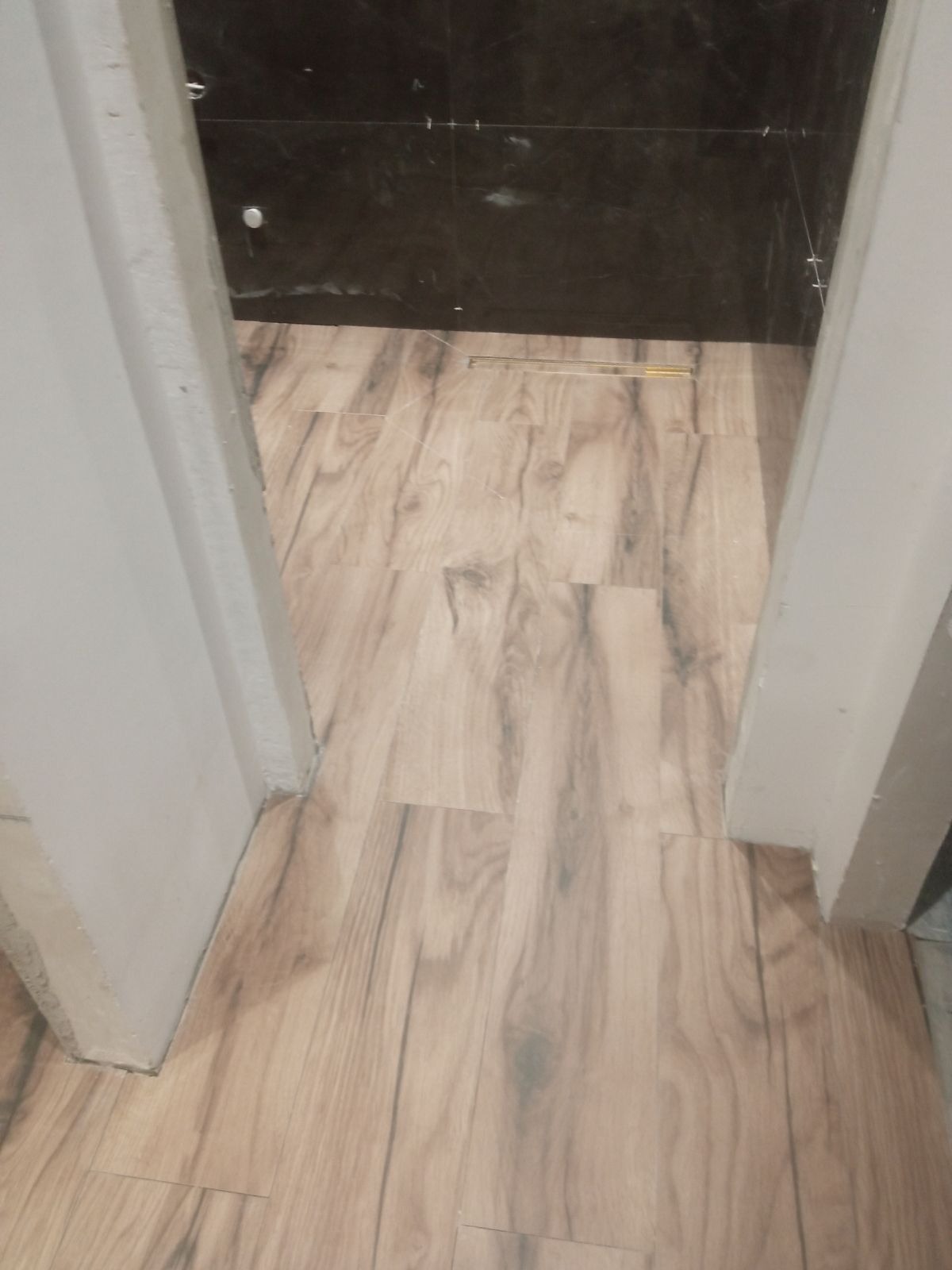 Ułożone płytki podłogowe imitujące drewno w łazience, widok zza progu, widoczny odpływ liniowy i fragment czarnej ściany z płytek