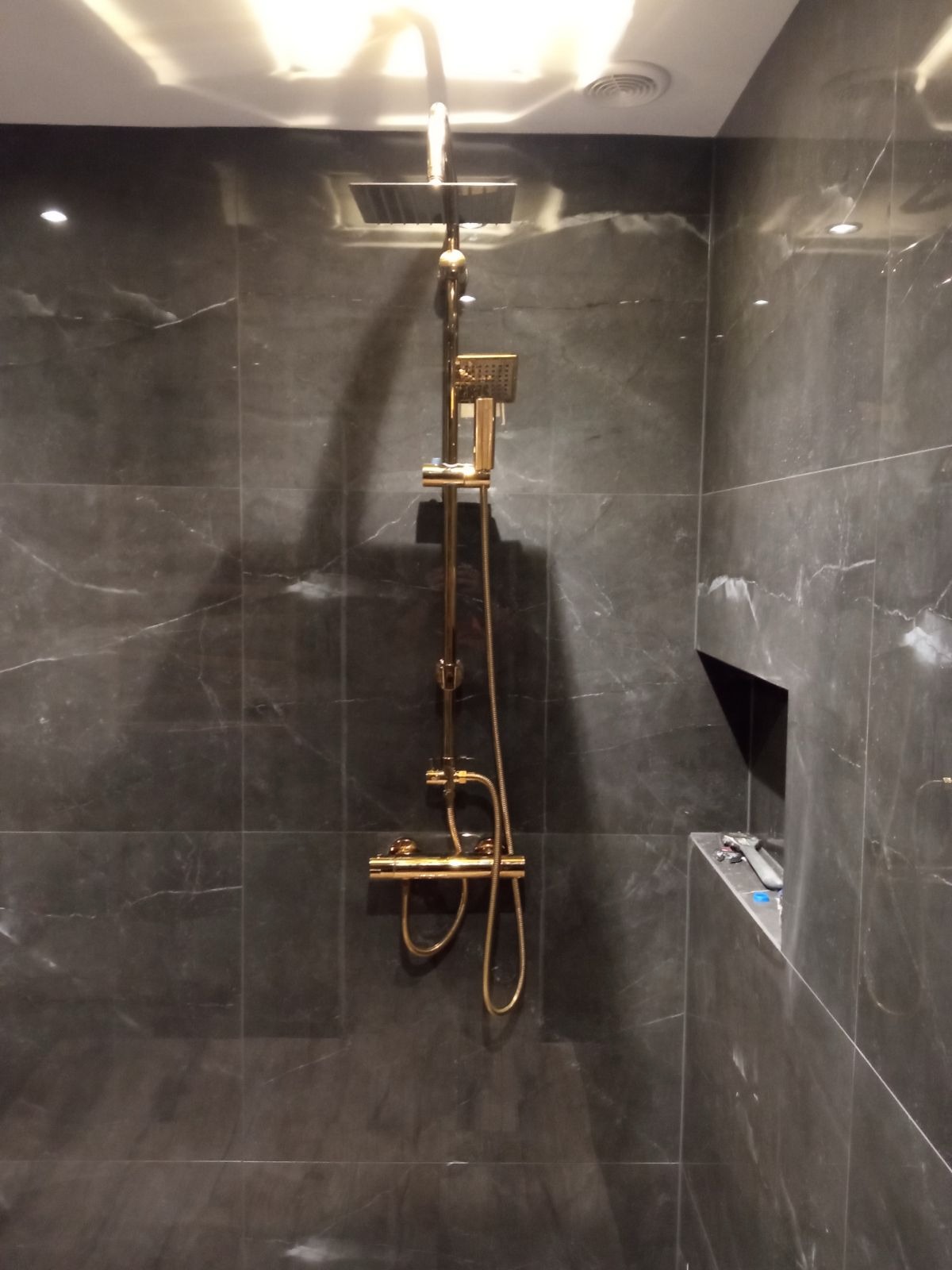 Wykończona łazienka z ciemnymi, marmurkowymi płytkami i złotą armaturą prysznicową; wnęka w ścianie z narzędziem glazurniczym.