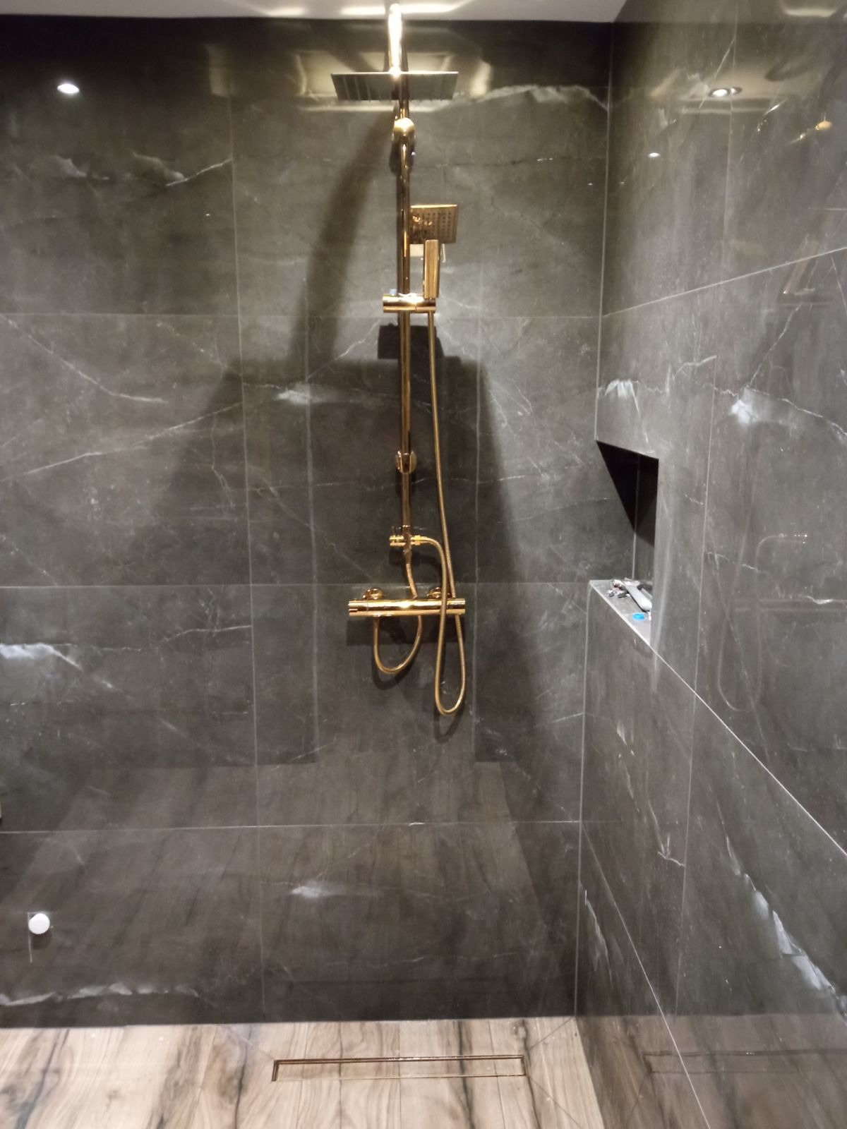 Nowoczesna kabina prysznicowa z armaturą w kolorze złotym i szarymi płytkami imitującymi marmur, odpływ liniowy w podłodze.