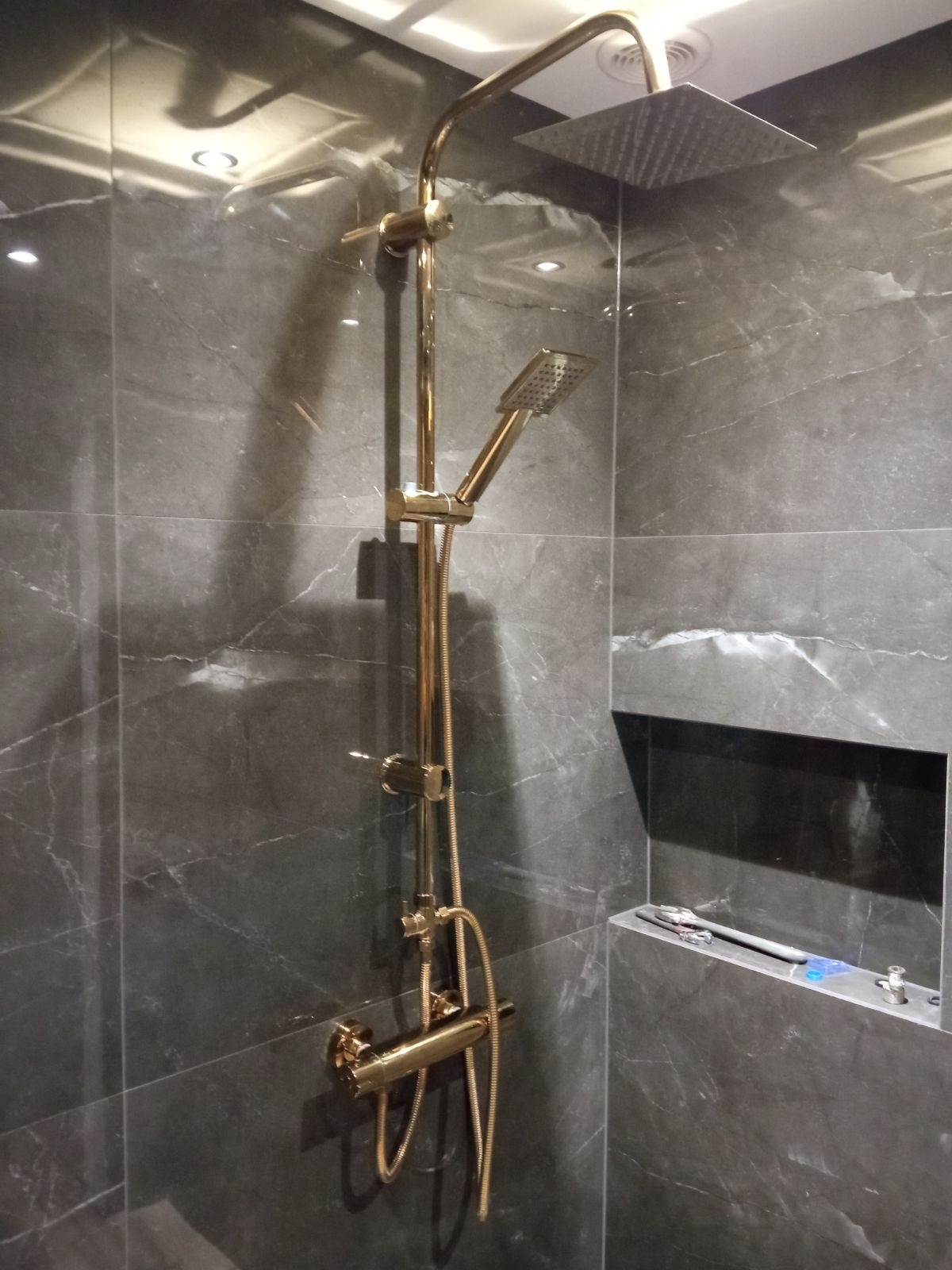 Złoty zestaw natryskowy z deszczownicą i słuchawką na tle szarych płytek imitujących marmur, widoczne narzędzia na półce, nowoczesna łazienka po remoncie.