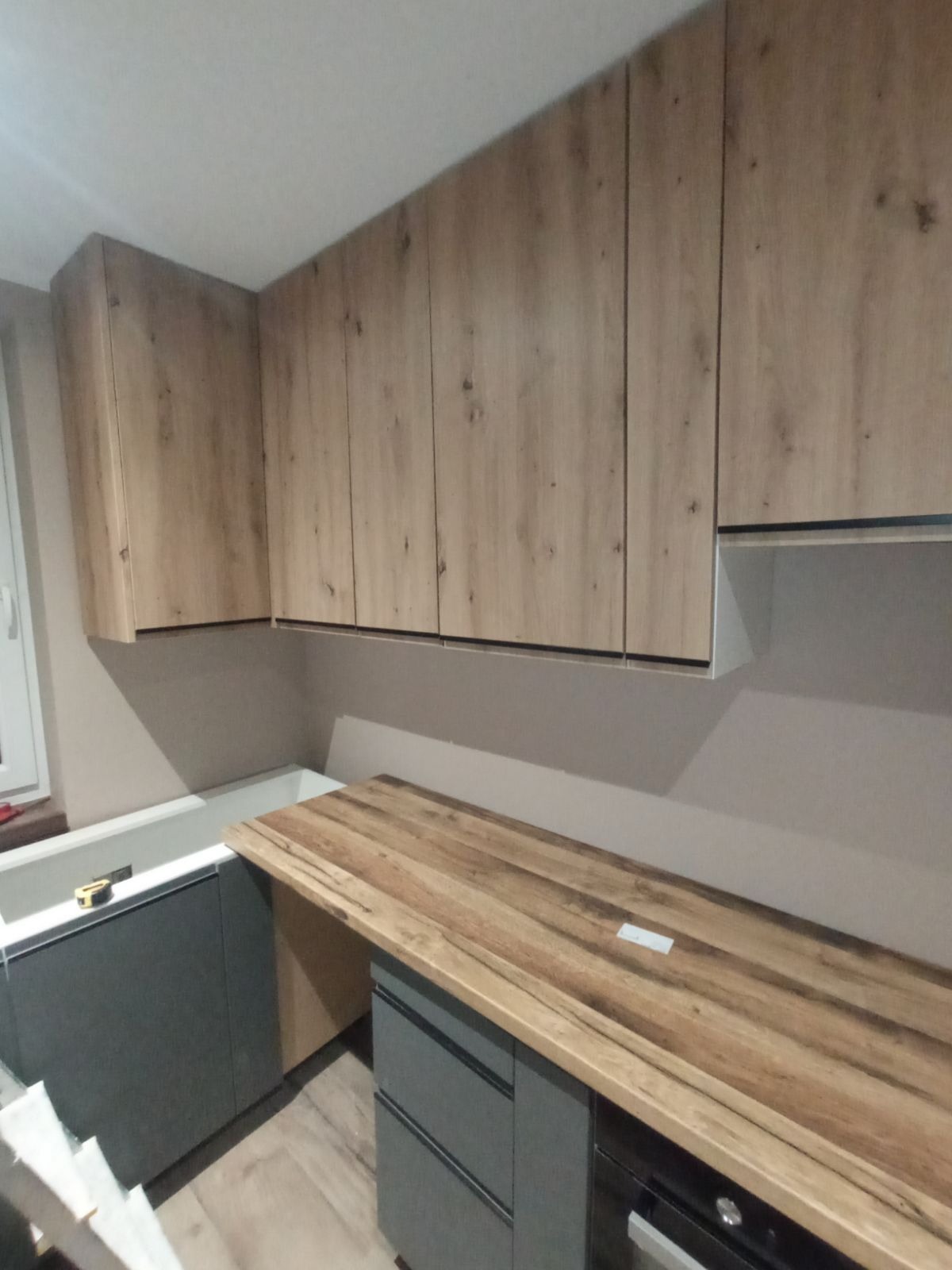 Nowoczesna kuchnia w trakcie montażu: drewnopodobne szafki wiszące i szare szafki dolne z drewnianym blatem, widoczne niedokończone wykończenie blatu.