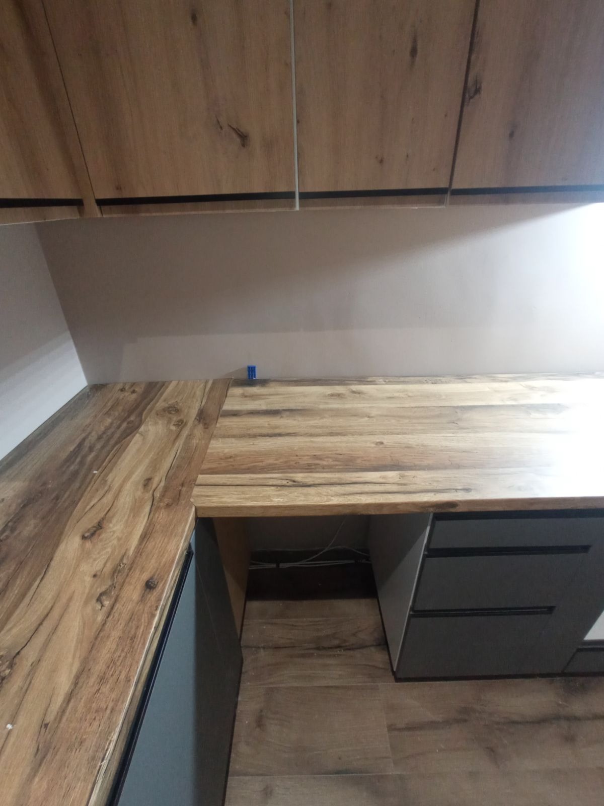 Nowoczesna kuchnia z drewnianymi szafkami górnymi i blatem w odcieniach brązu, szare szafki dolne z szufladami, widoczne okablowanie pod blatem, niebieski element mocujący na blacie.