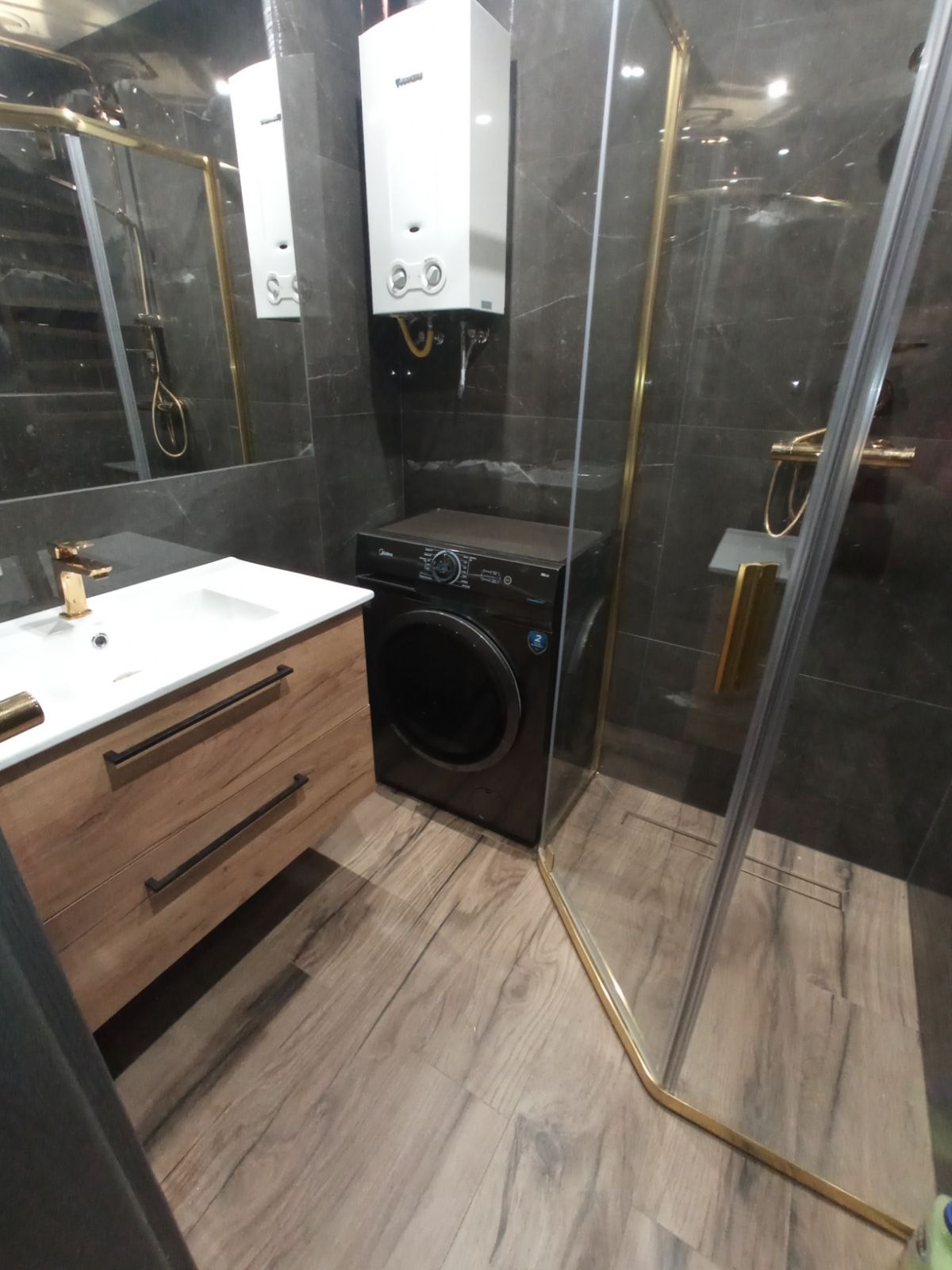 Nowoczesna łazienka z czarną pralką automatyczną, umywalką z drewnianą szafką, kabiną prysznicową ze złotymi ramami i szarą marmurową ścianą, oświetlona światłem z góry.