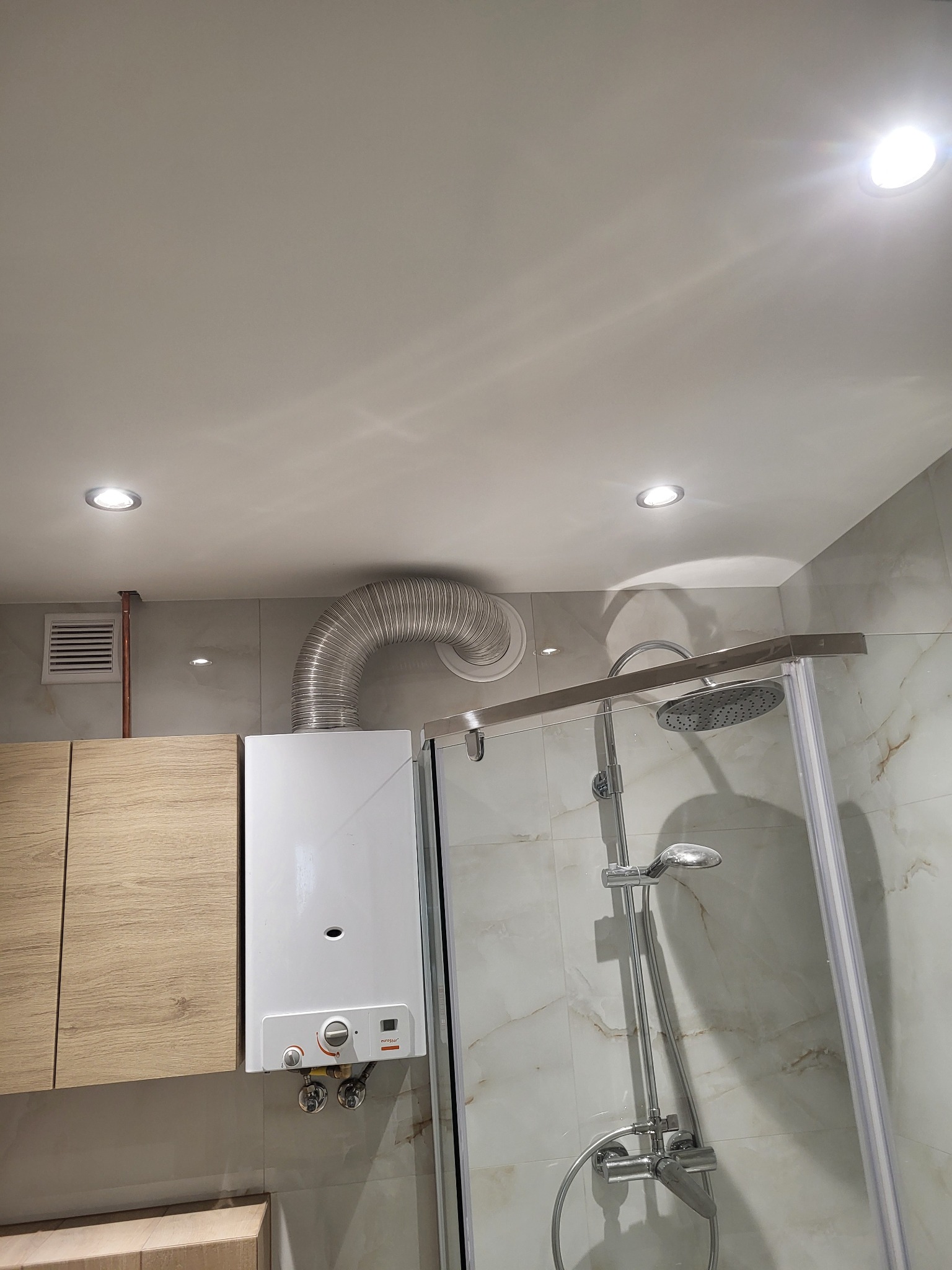 Biały piec gazowy z podłączoną aluminiową rurą wentylacyjną w łazience z kabiną prysznicową, zamontowany obok drewnianych szafek, oświetlony punktowymi lampami sufitowymi.