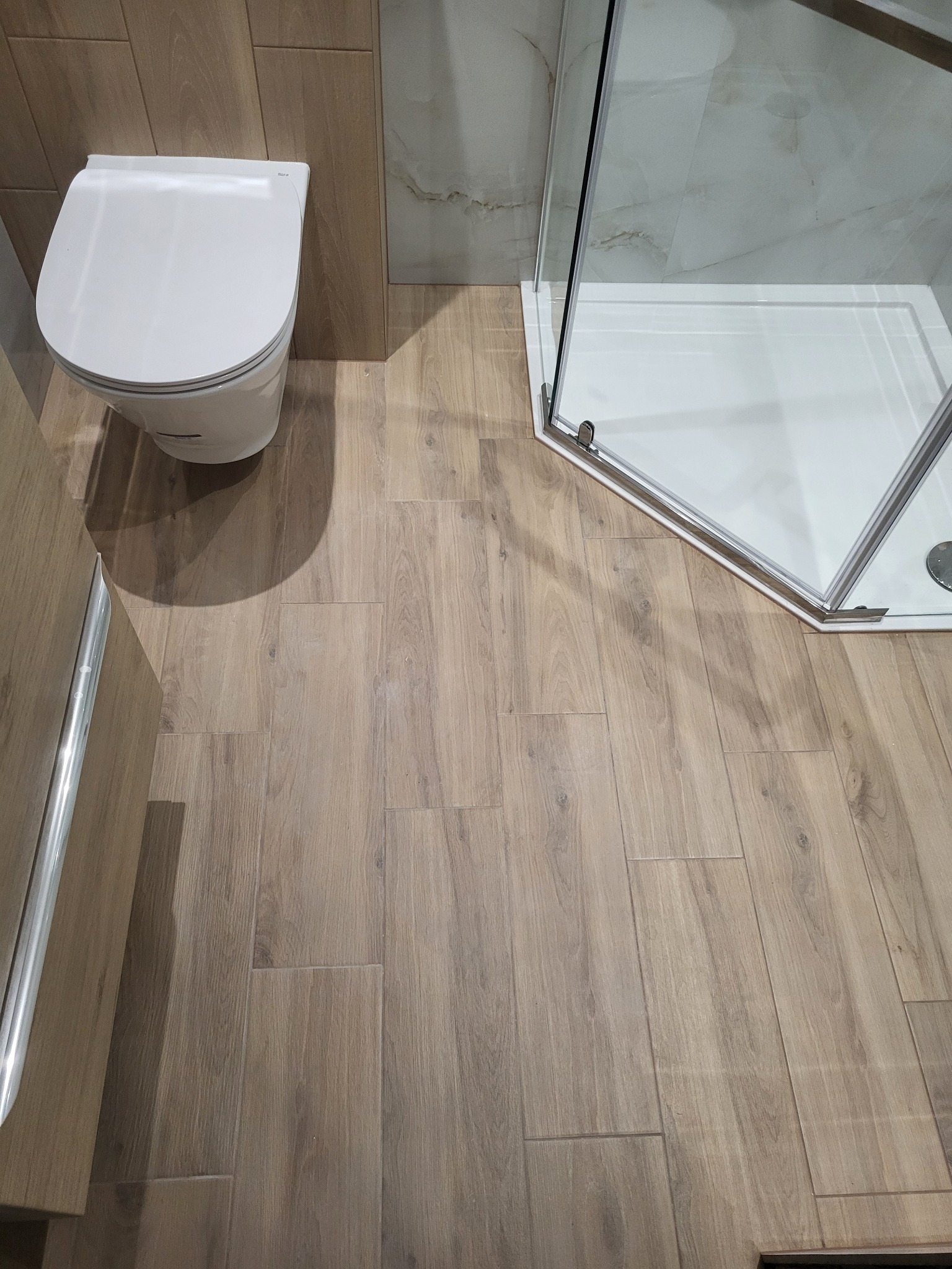 Wykończona łazienka z kabiną prysznicową i toaletą, drewnopodobne płytki podłogowe ułożone w prosty wzór, jasne ściany częściowo wykończone płytkami imitującymi marmur.