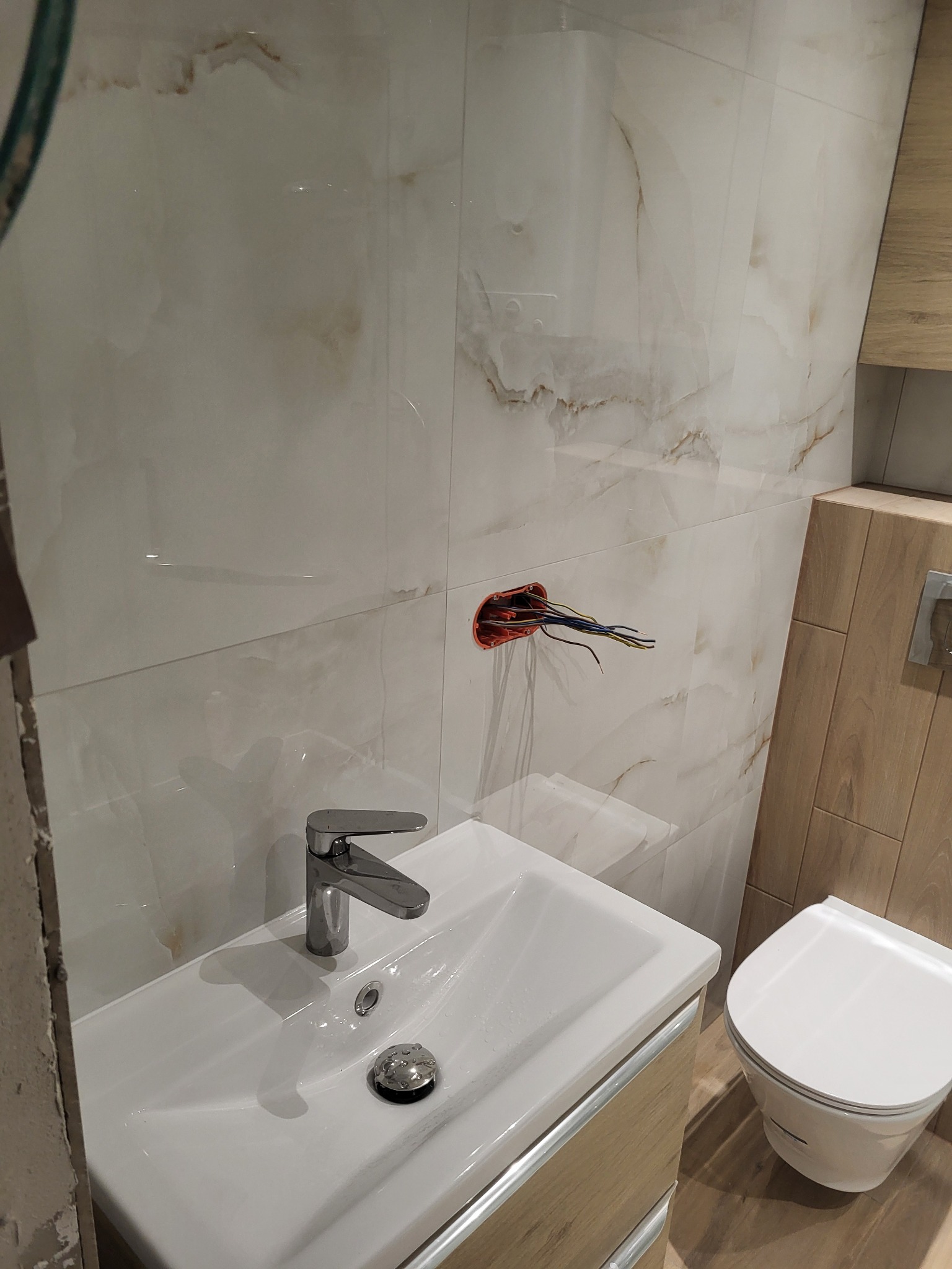 Łazienka w trakcie prac wykończeniowych: zamontowana umywalka z chromowaną baterią, widoczne wyprowadzone przewody elektryczne ze ściany wyłożonej jasnymi płytkami imitującymi marmur, częściowo...