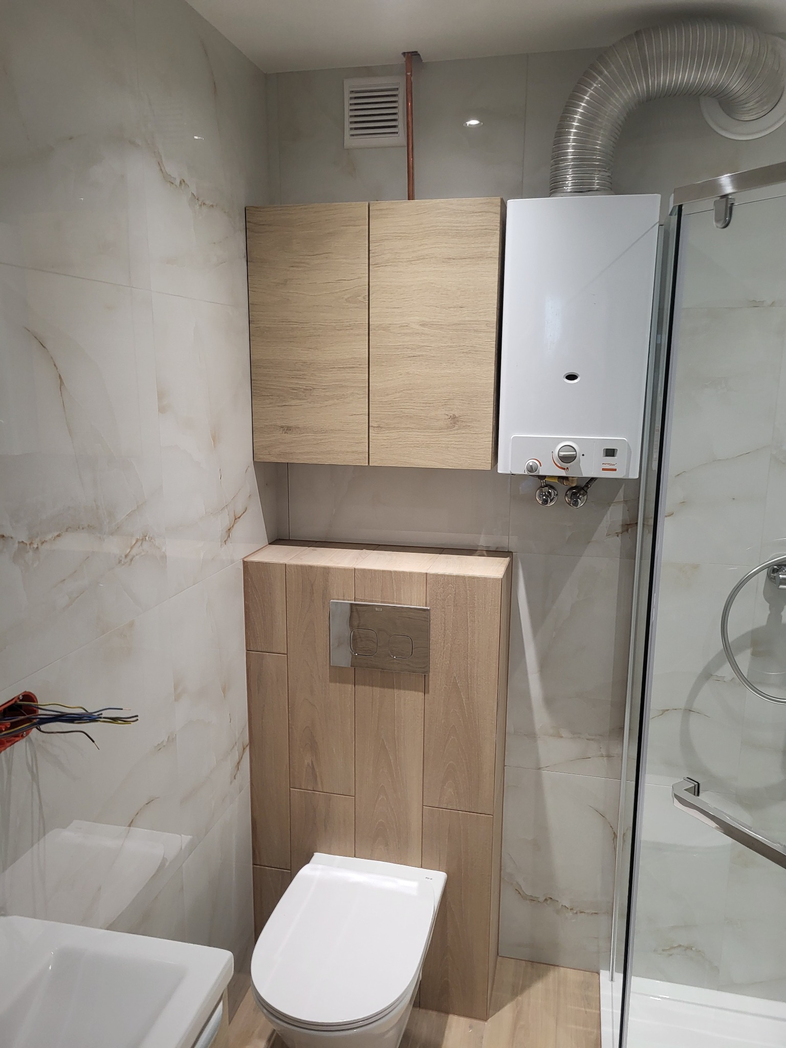 Wykończona łazienka z wiszącą szafką, zabudową WC z przyciskiem Roca, białym piecem gazowym z podłączeniem do komina oraz kabiną prysznicową z elementami chromowanymi, wykończona płytkami...