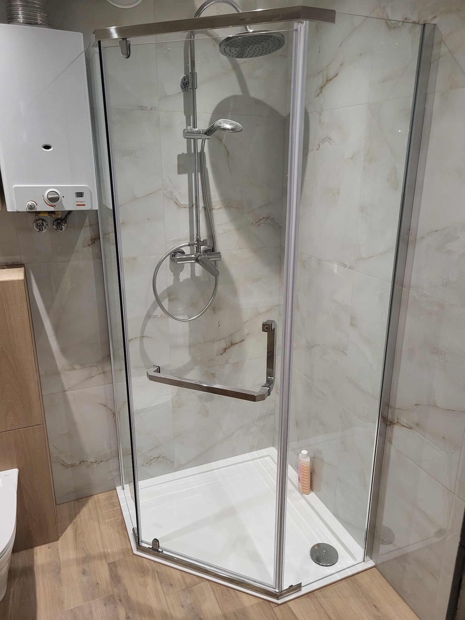 Narożna kabina prysznicowa z chromowanymi profilami i deszczownicą, widoczny podgrzewacz wody i marmurowe płytki ścienne.