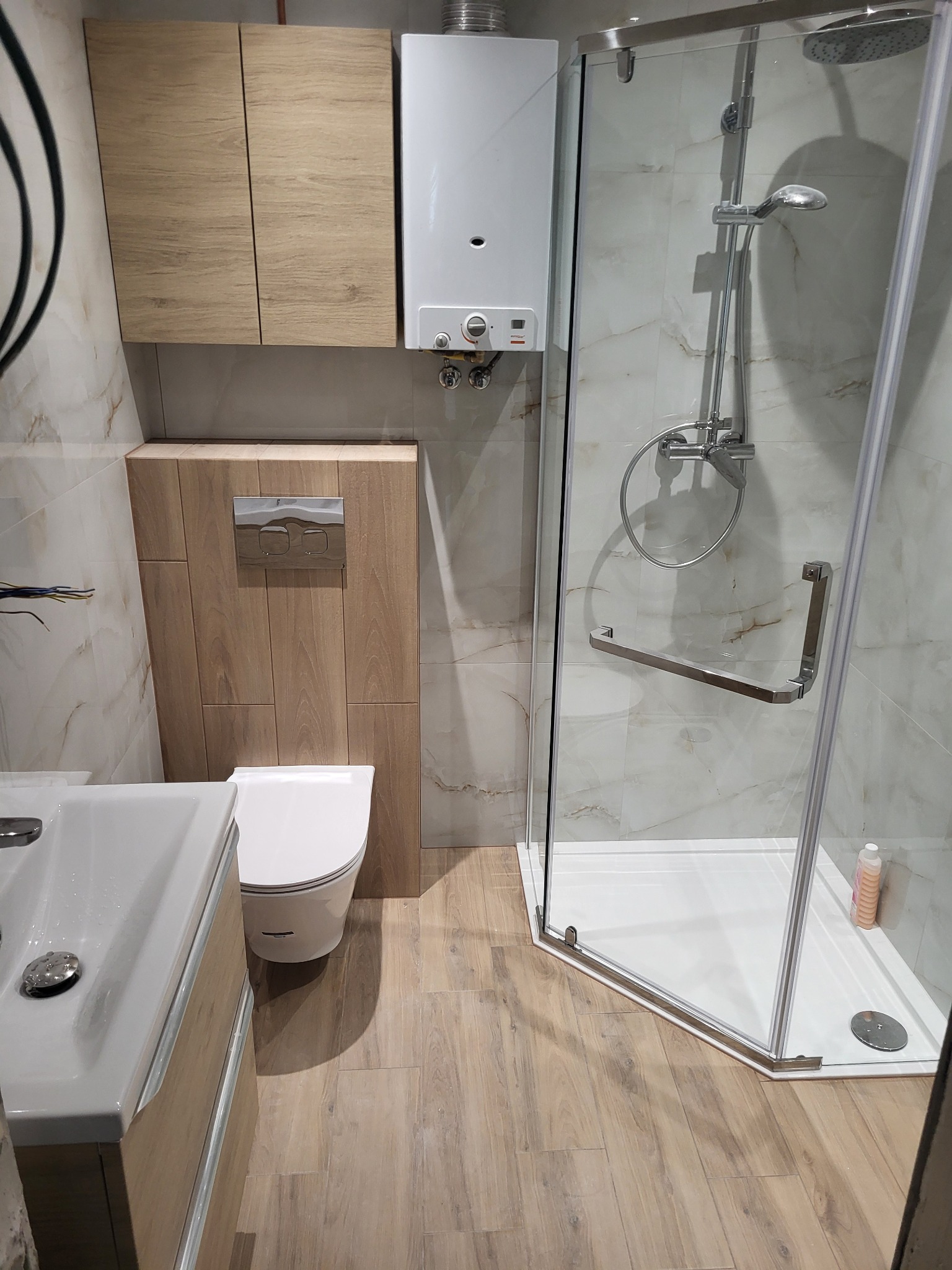 Nowoczesna łazienka po remoncie z kabiną prysznicową narożną, wiszącą toaletą, szafkami z drewnopodobnym frontem oraz piecykiem gazowym nad zabudową toalety.