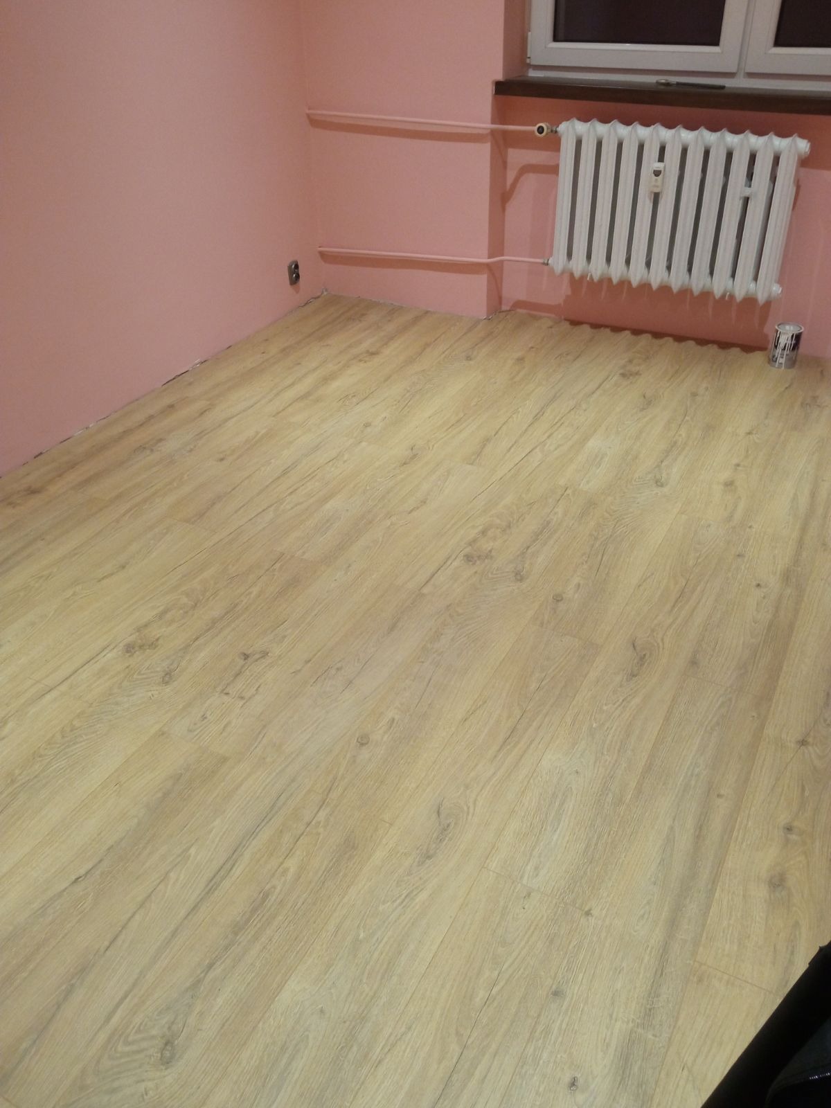 Ułożone panele podłogowe w pokoju z różowymi ścianami i białym grzejnikiem, widoczna puszka po farbie przy grzejniku.