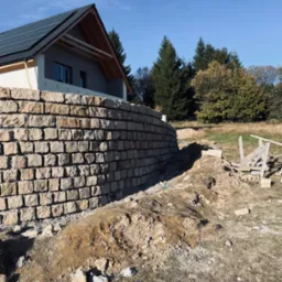 Murowana ściana oporowa z kamienia naturalnego przy nowo wybudowanym domu z panelami słonecznymi na dachu, widoczne ślady prac ziemnych i drewniane rusztowanie.