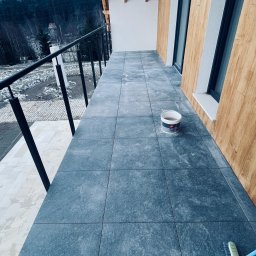 Construction Beton and Steel Poland - Wykończony balkon z szarymi płytkami, widok na zalesione wzgórza w pochmurny dzień, wiadro z preparatem do czyszczenia płytek i szczotka do zamiatania.