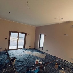 Construction Beton and Steel Poland - Pokój w trakcie prac wykończeniowych: świeżo pomalowane ściany w odcieniach beżu, podłoga zabezpieczona folią malarską, widoczne puszki elektryczne i wystające przewody, okno balkonowe z widokiem...