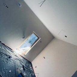 Construction Beton and Steel Poland - Poddasze w trakcie prac wykończeniowych: widoczne okno dachowe, zabezpieczona folią ściana, surowe ściany z widocznymi punktami elektrycznymi i wystającymi kablami.