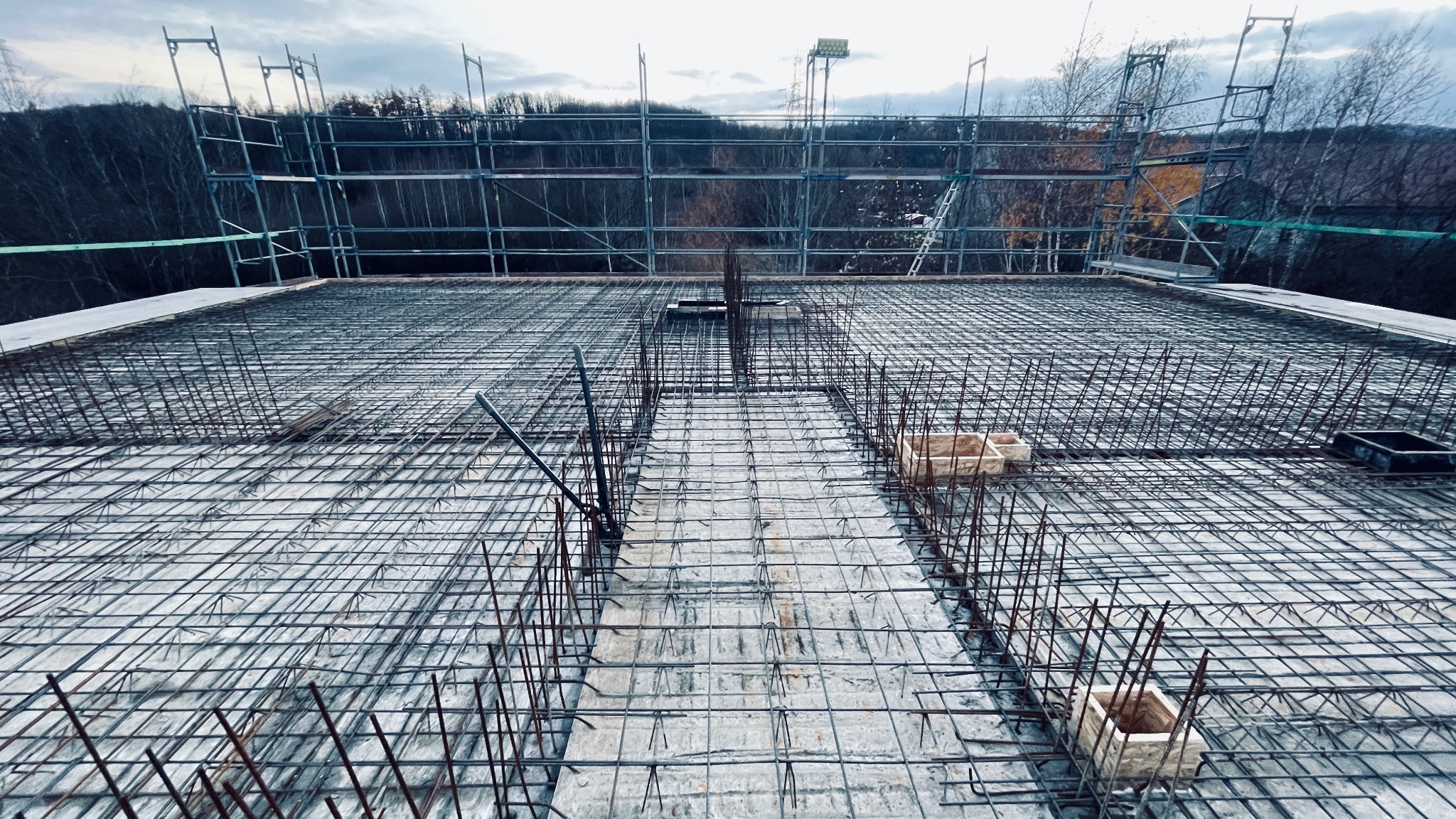 Zbrojenie płyty fundamentowej z widocznymi prętami stalowymi, siatką zbrojeniową i drewnianymi elementami szalunku, rusztowanie w tle.