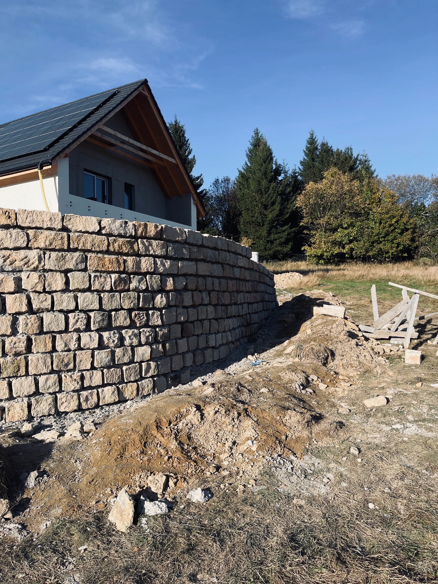 Murowana ściana oporowa z kamienia naturalnego przy nowo wybudowanym domu z panelami słonecznymi na dachu, widoczne ślady prac ziemnych i drewniane rusztowanie.