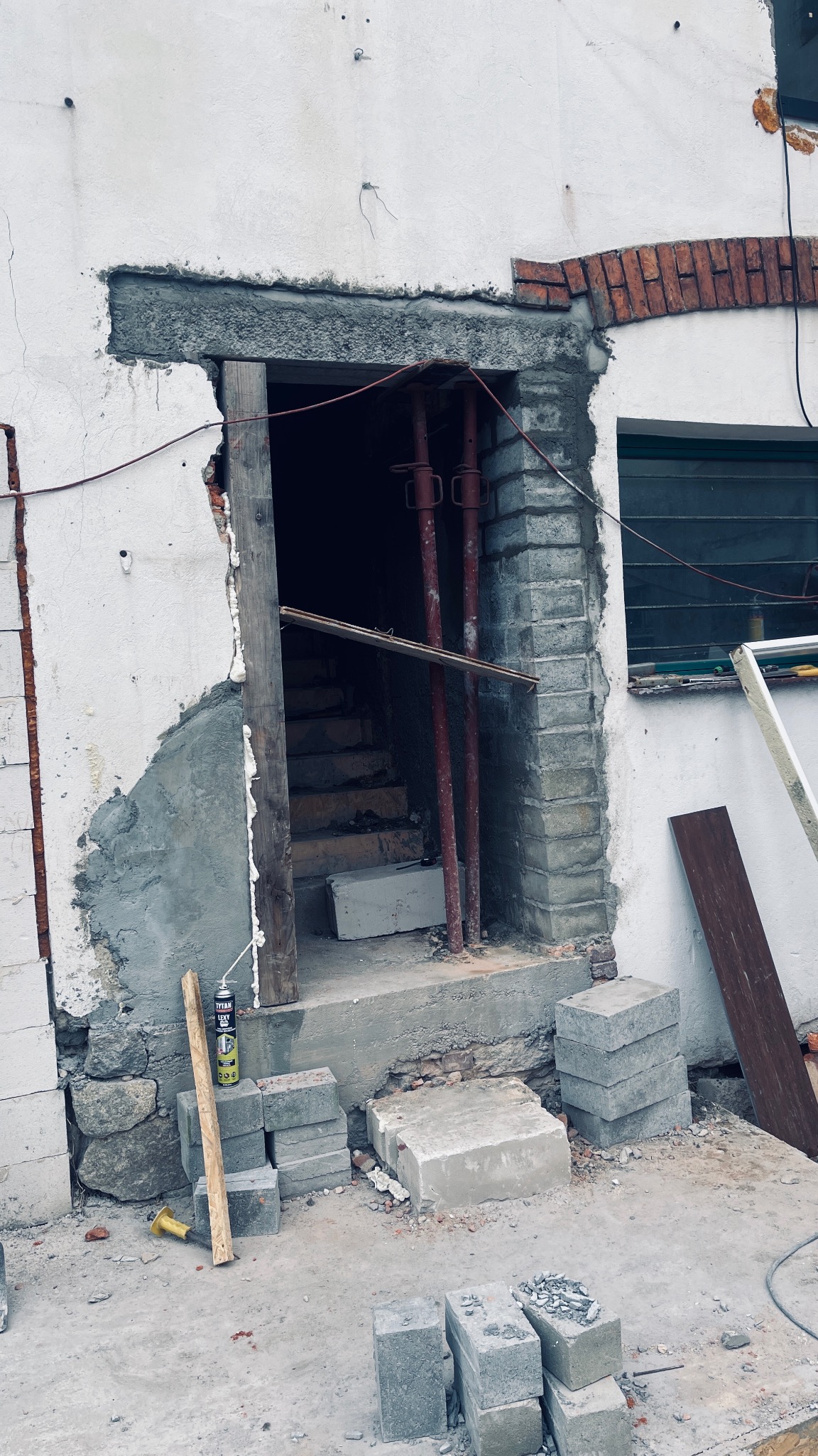 Otwór drzwiowy w trakcie murowania z widocznymi cegłami, pustakami, podporami stalowymi, pianką montażową i schodami w głębi.