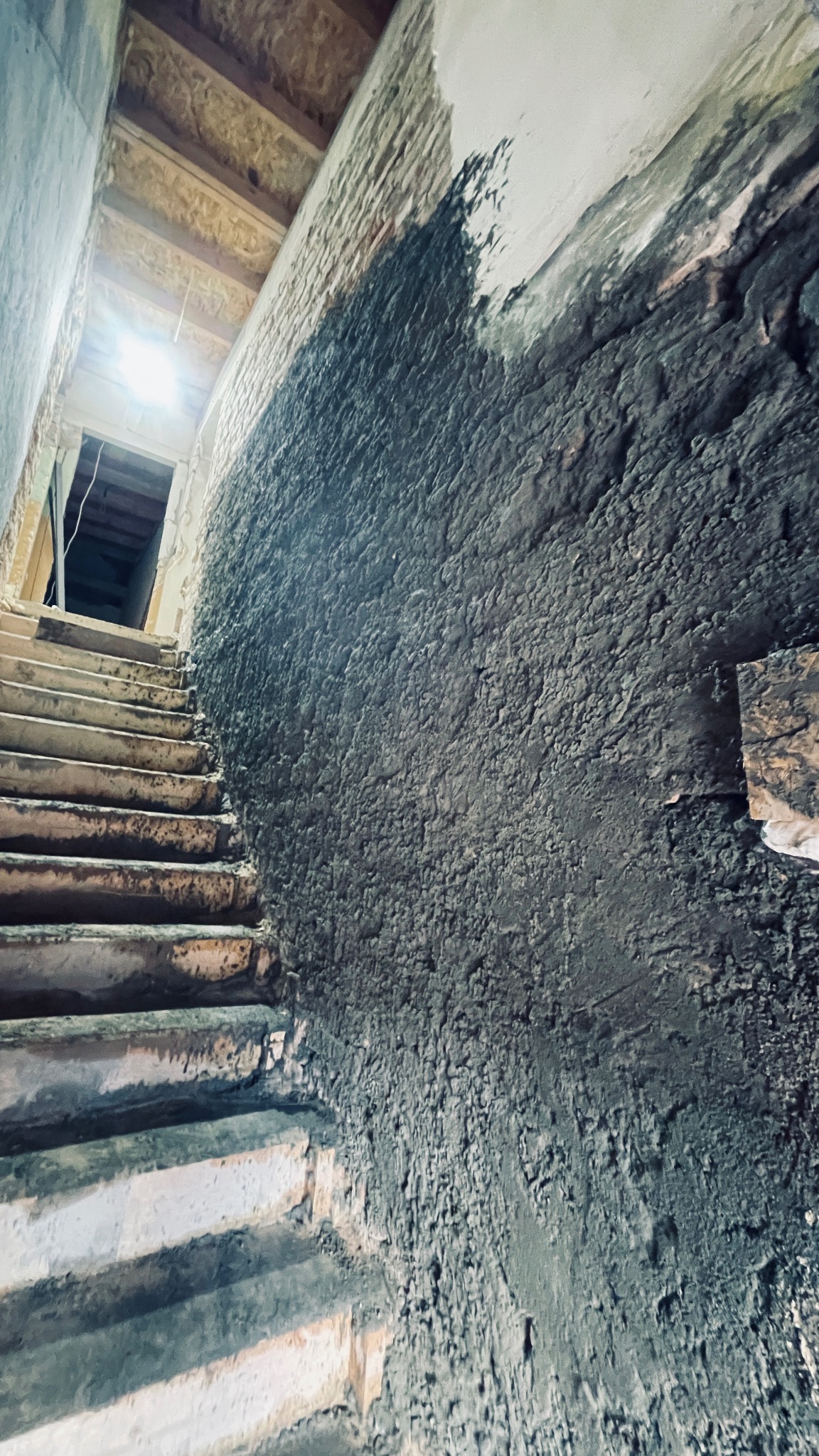 Surowe, betonowe schody prowadzące w górę, obok ściany z nierówną, szarą fakturą, częściowo pokrytą świeżym tynkiem.