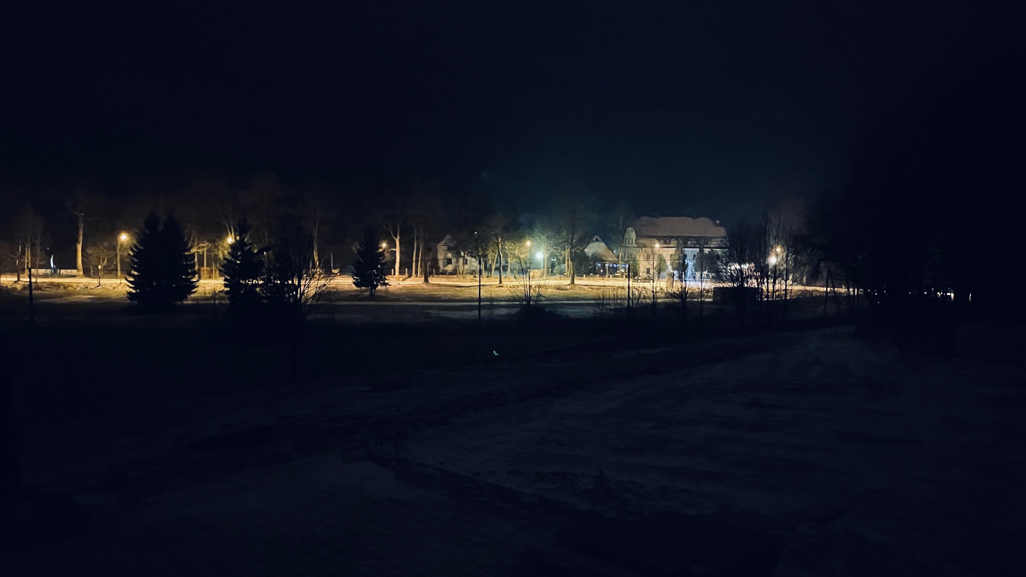 Nocny widok na oświetlony budynek z czerwonym dachem, otoczony drzewami i śniegiem, oświetlony latarniami.