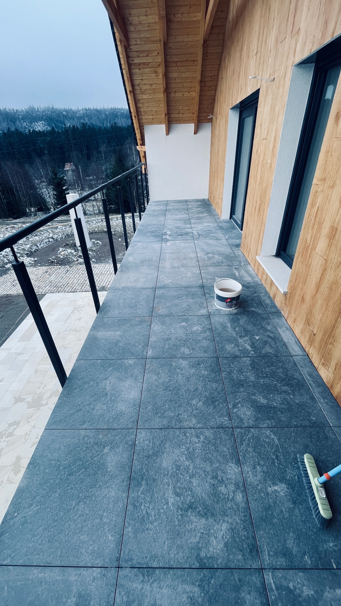 Wykończony balkon z szarymi płytkami, widok na zalesione wzgórza w pochmurny dzień, wiadro z preparatem do czyszczenia płytek i szczotka do zamiatania.