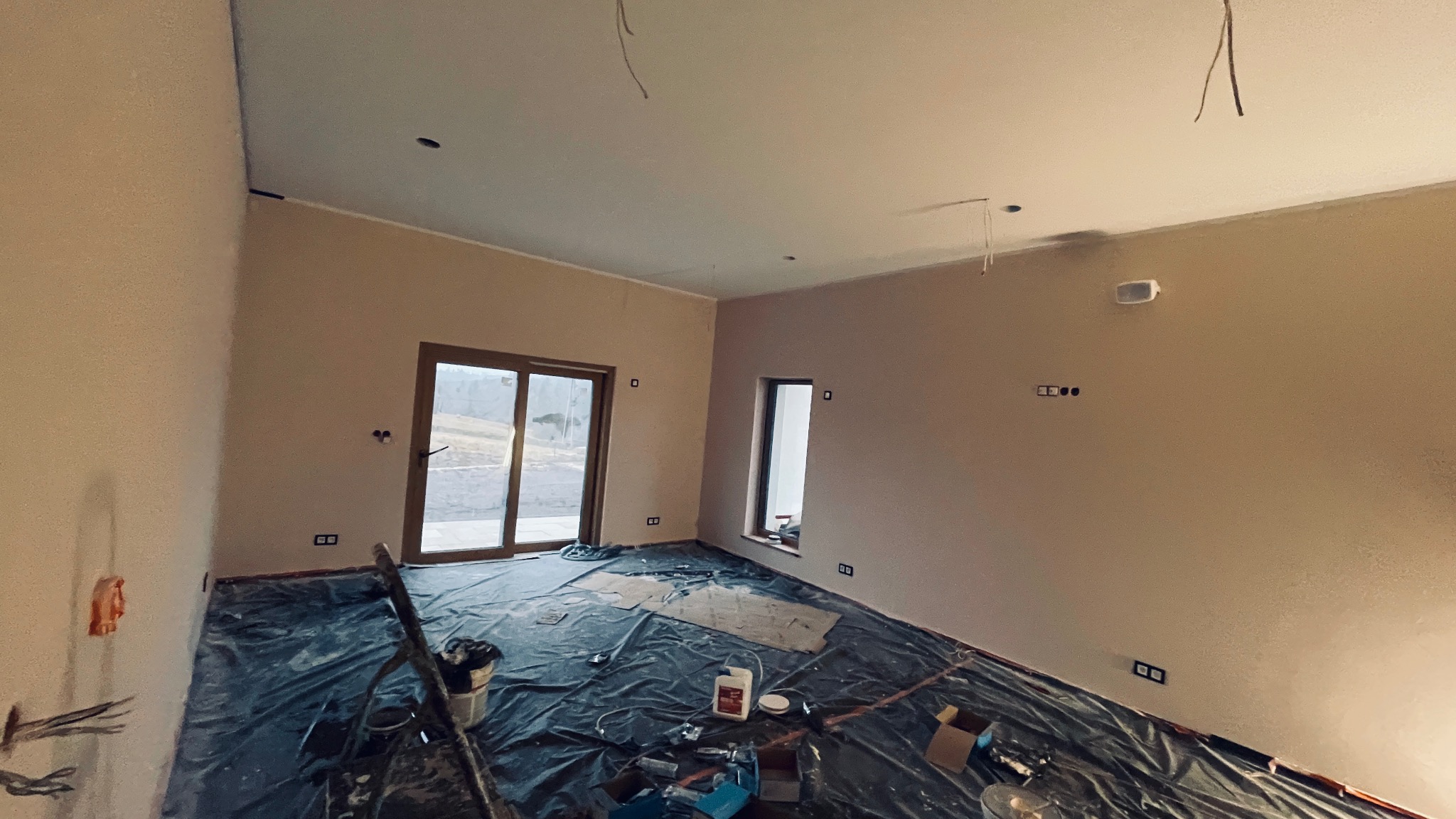 Pokój w trakcie prac wykończeniowych: świeżo pomalowane ściany w odcieniach beżu, podłoga zabezpieczona folią malarską, widoczne puszki elektryczne i wystające przewody, okno balkonowe z widokiem...