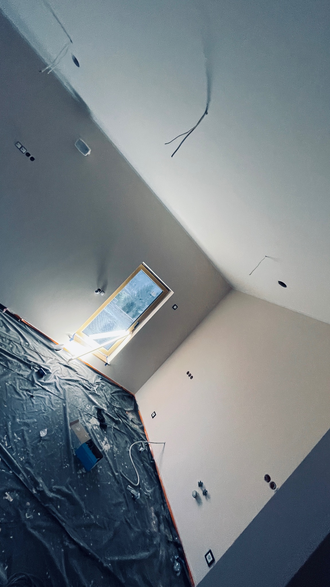 Poddasze w trakcie prac wykończeniowych: widoczne okno dachowe, zabezpieczona folią ściana, surowe ściany z widocznymi punktami elektrycznymi i wystającymi kablami.