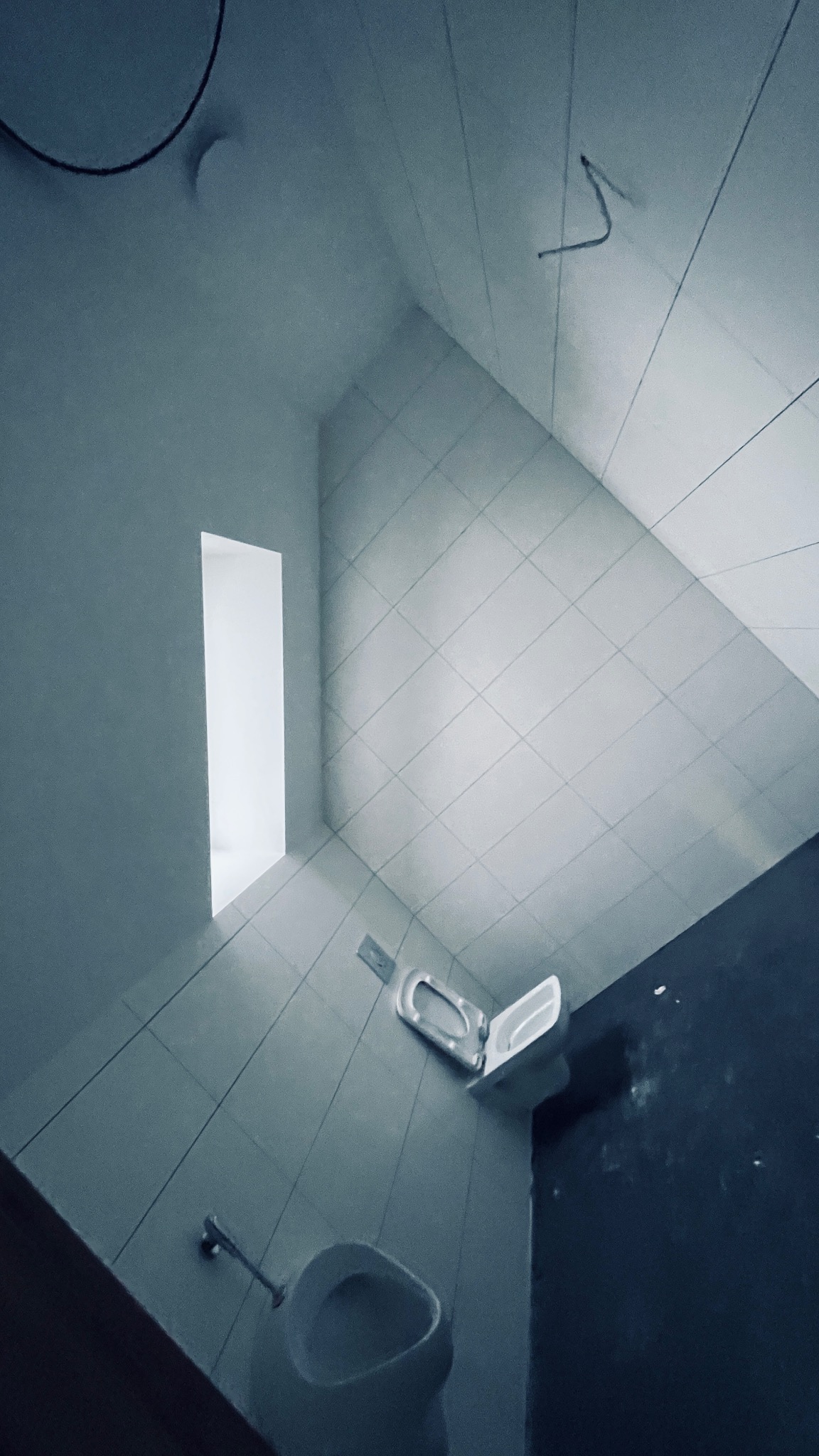 Wnętrze łazienki w trakcie remontu, widok od dołu: białe płytki na ścianach i podłodze, częściowo zamontowana toaleta, otwór okienny bez okna, widoczne ślady po pracach wykończeniowych.