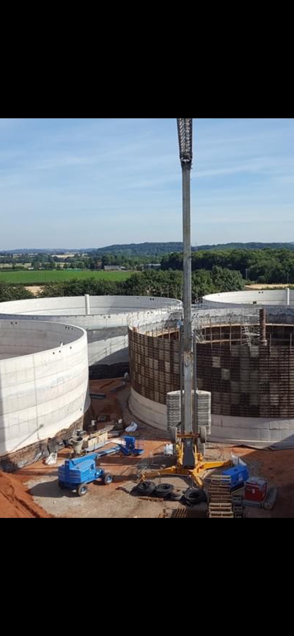 Wysoki dźwig na placu budowy trzech okrągłych zbiorników z betonu, widok z góry na tle zielonych pól i lasów.