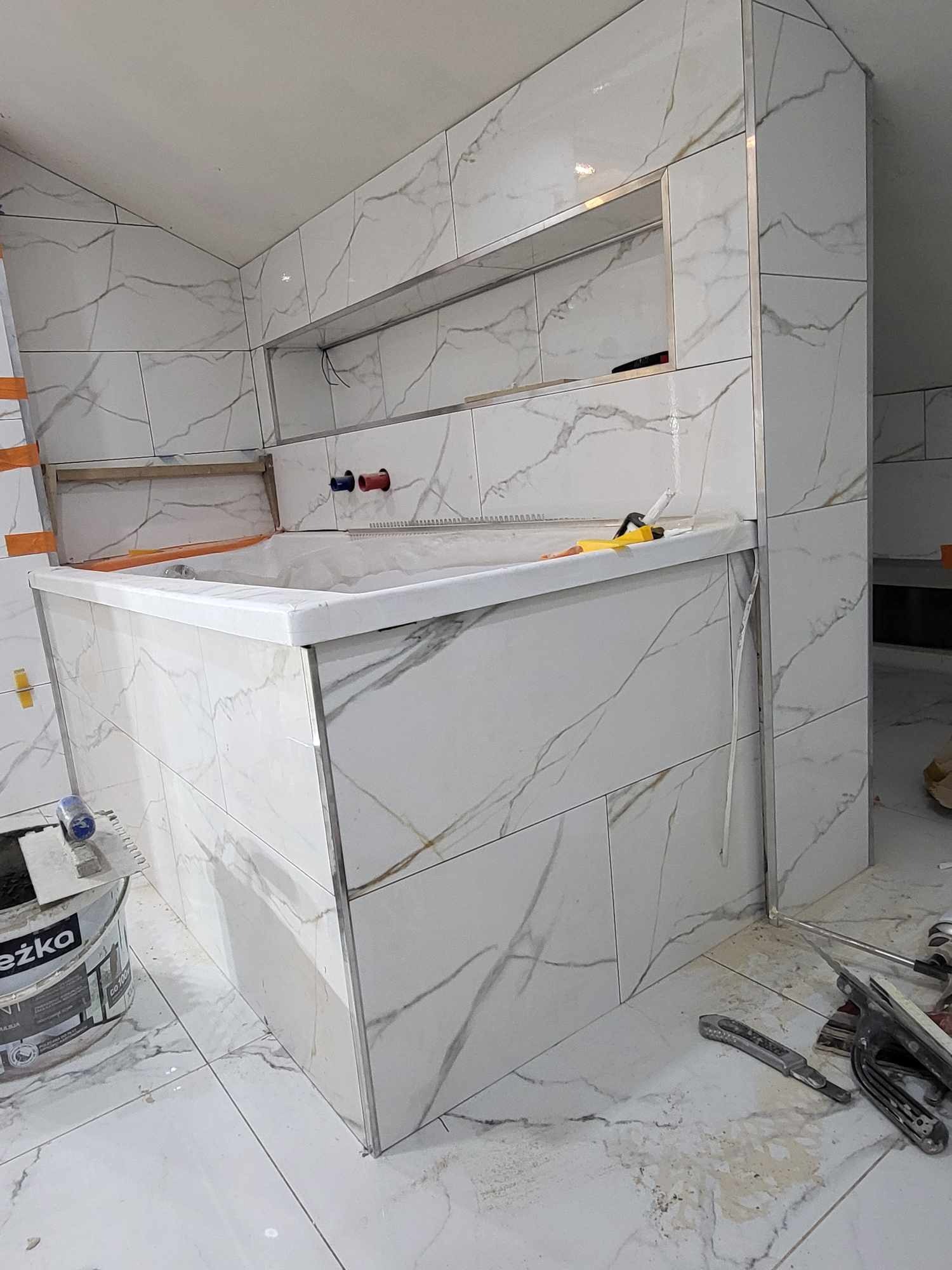 Łazienka w trakcie remontu: obudowa wanny i ściany wyłożone białymi płytkami imitującymi marmur, widoczne narzędzia glazurnicze i wiadro z klejem, surowe wykończenie.