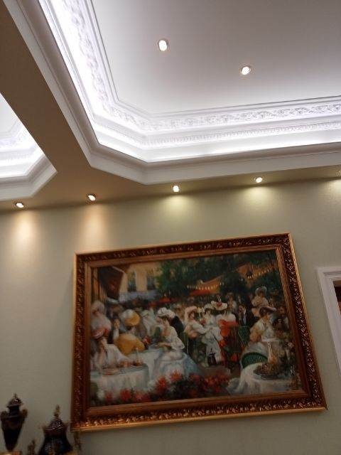 Oświetlenie sufitowe LED z dekoracyjnymi sztukateriami i obrazem w złotej ramie na ścianie w kolorze ecru.