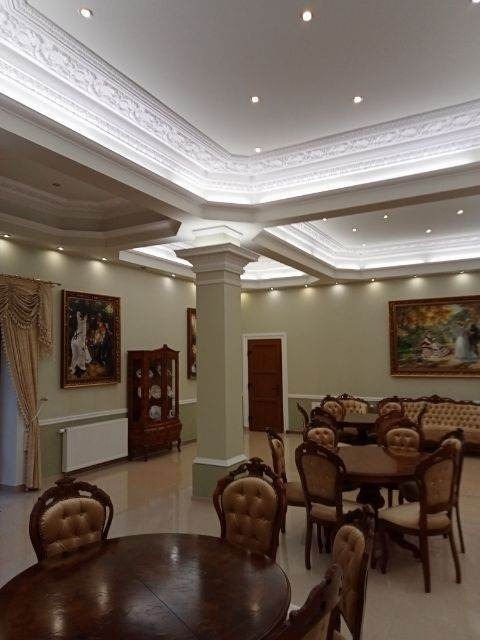 Eleganckie wnętrze sali z dekoracyjnym sufitem podwieszanym z oświetleniem punktowym, kolumną architektoniczną, klasycznymi meblami i obrazami w ozdobnych ramach.