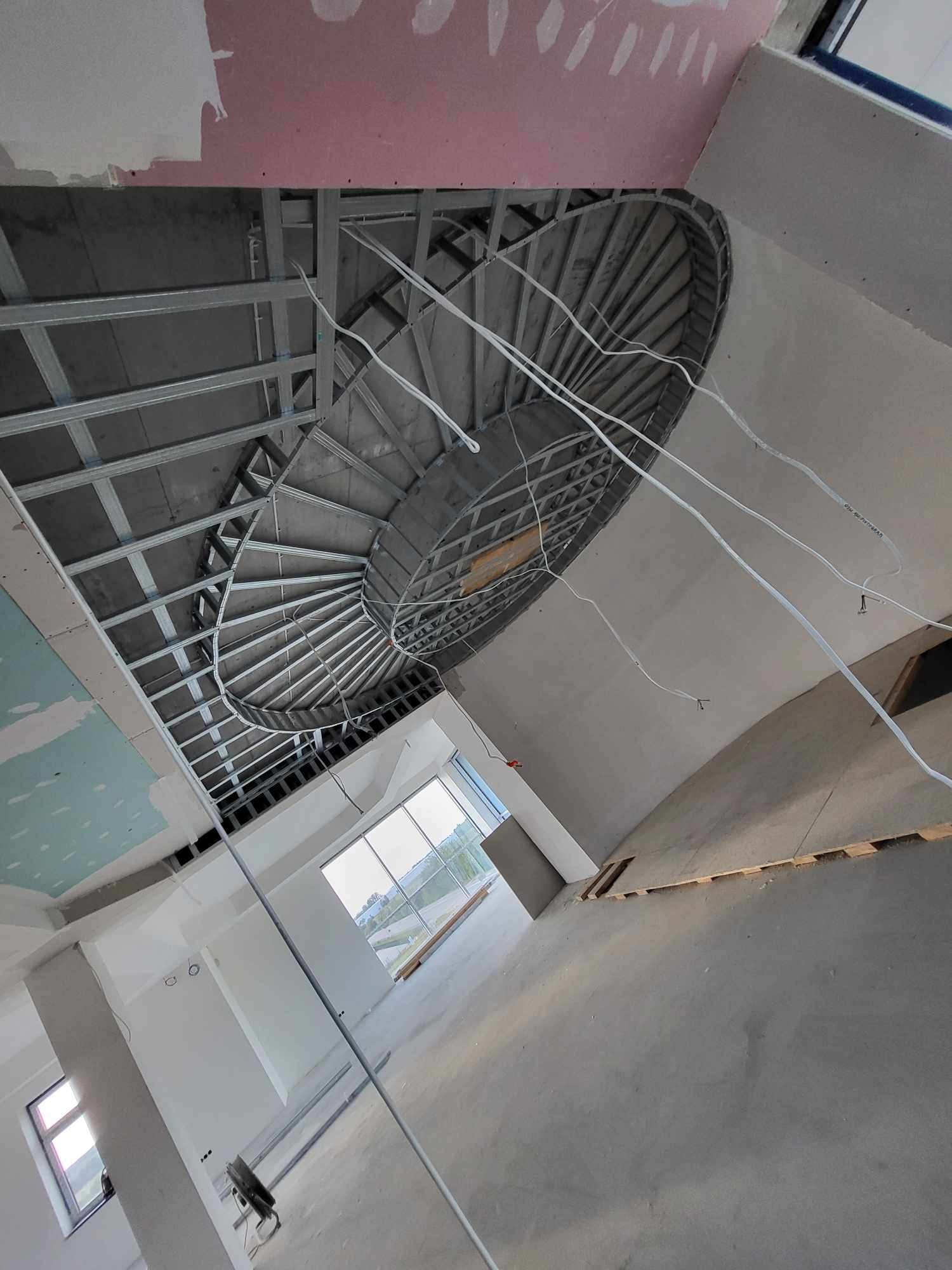 Wnętrze w stanie surowym otwartym, widok na sufit z metalową konstrukcją podwieszaną i okrągłym otworem, wystające kable, ściany częściowo pokryte płytami gipsowo-kartonowymi, duże okna w tle.