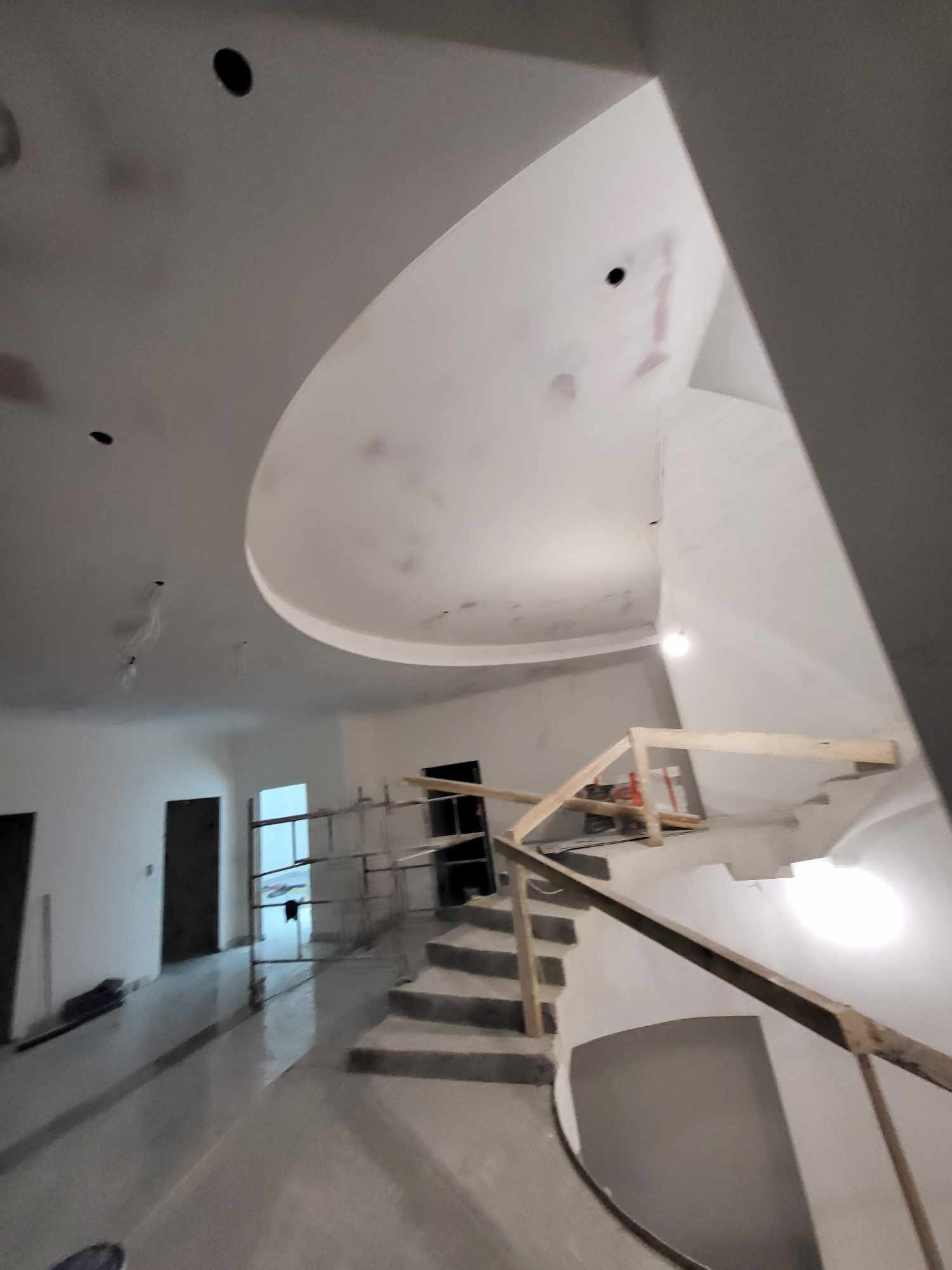 Wnętrze w trakcie prac wykończeniowych: surowa klatka schodowa z drewnianą barierką i schodami z betonu, widoczne rusztowania i niedokończony sufit z owalnym wgłębieniem, ściany przygotowane...