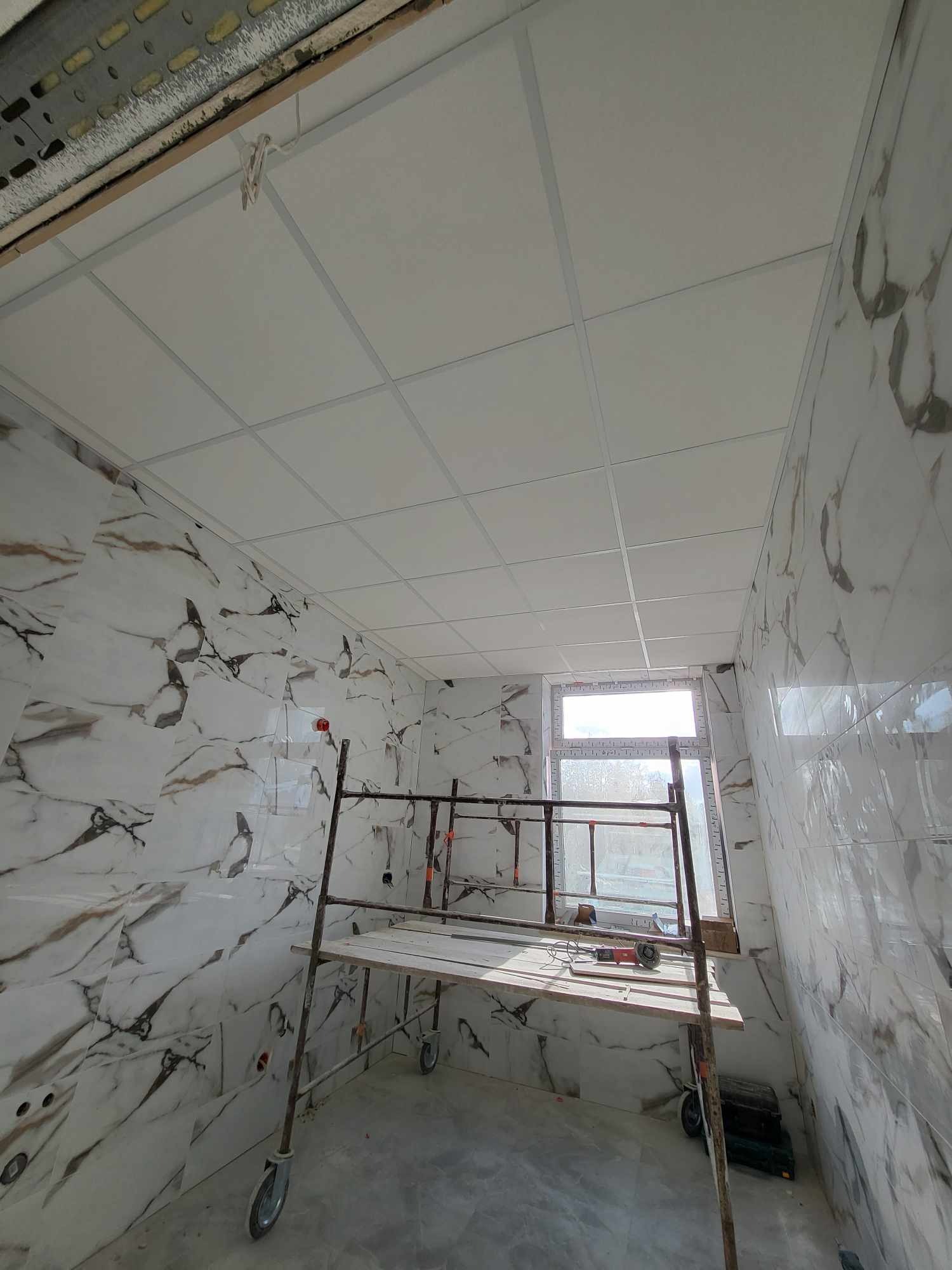Łazienka w trakcie montażu sufitu podwieszanego z płyt kwadratowych, ściany wyłożone płytkami imitującymi marmur, rusztowanie robocze w pomieszczeniu.