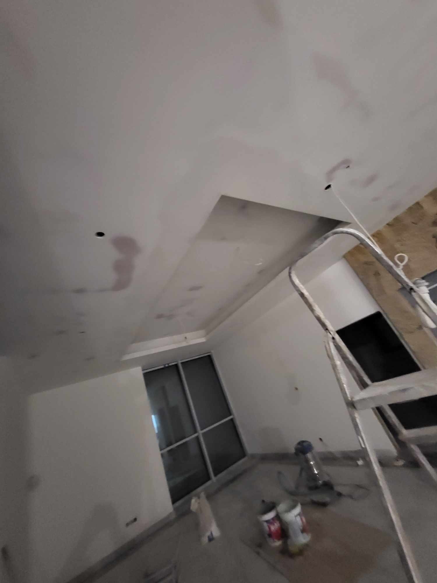 Przygotowanie sufitu do malowania: widoczne ślady po szpachlowaniu, drabina aluminiowa, puszki z farbą i szlifierka na podłodze, okno w tle.