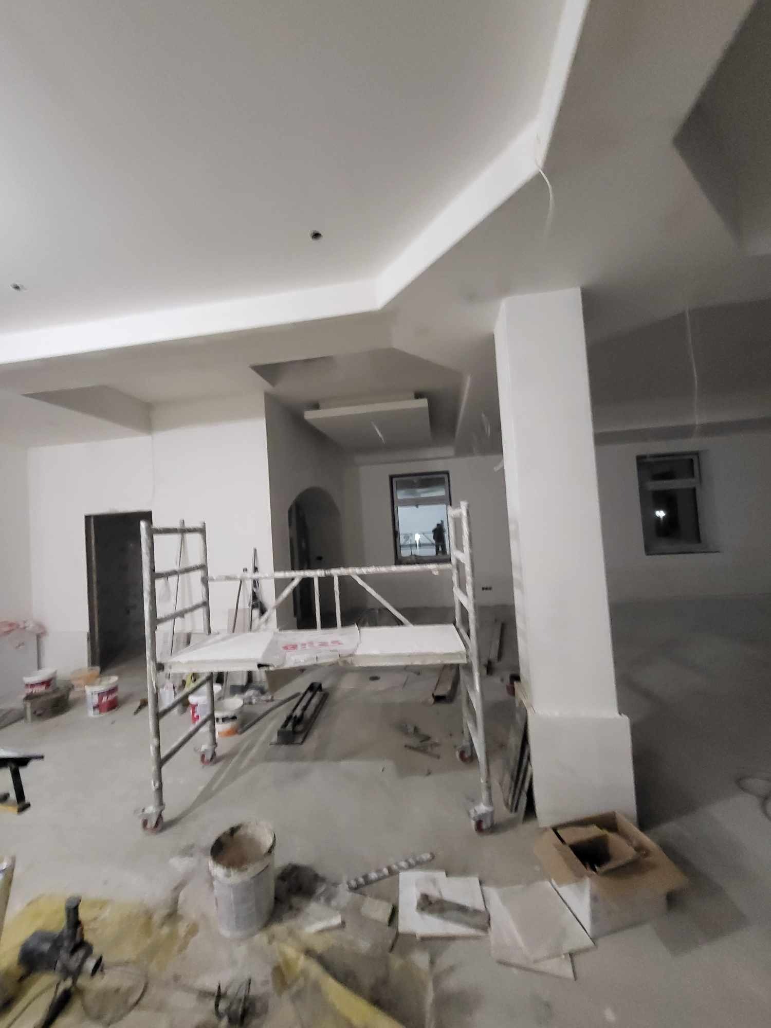 Wnętrze w trakcie prac wykończeniowych; widoczne rusztowanie, puszki z farbą, narzędzia malarskie i płytki podłogowe, świeżo pomalowane ściany i sufit z elementami dekoracyjnymi.