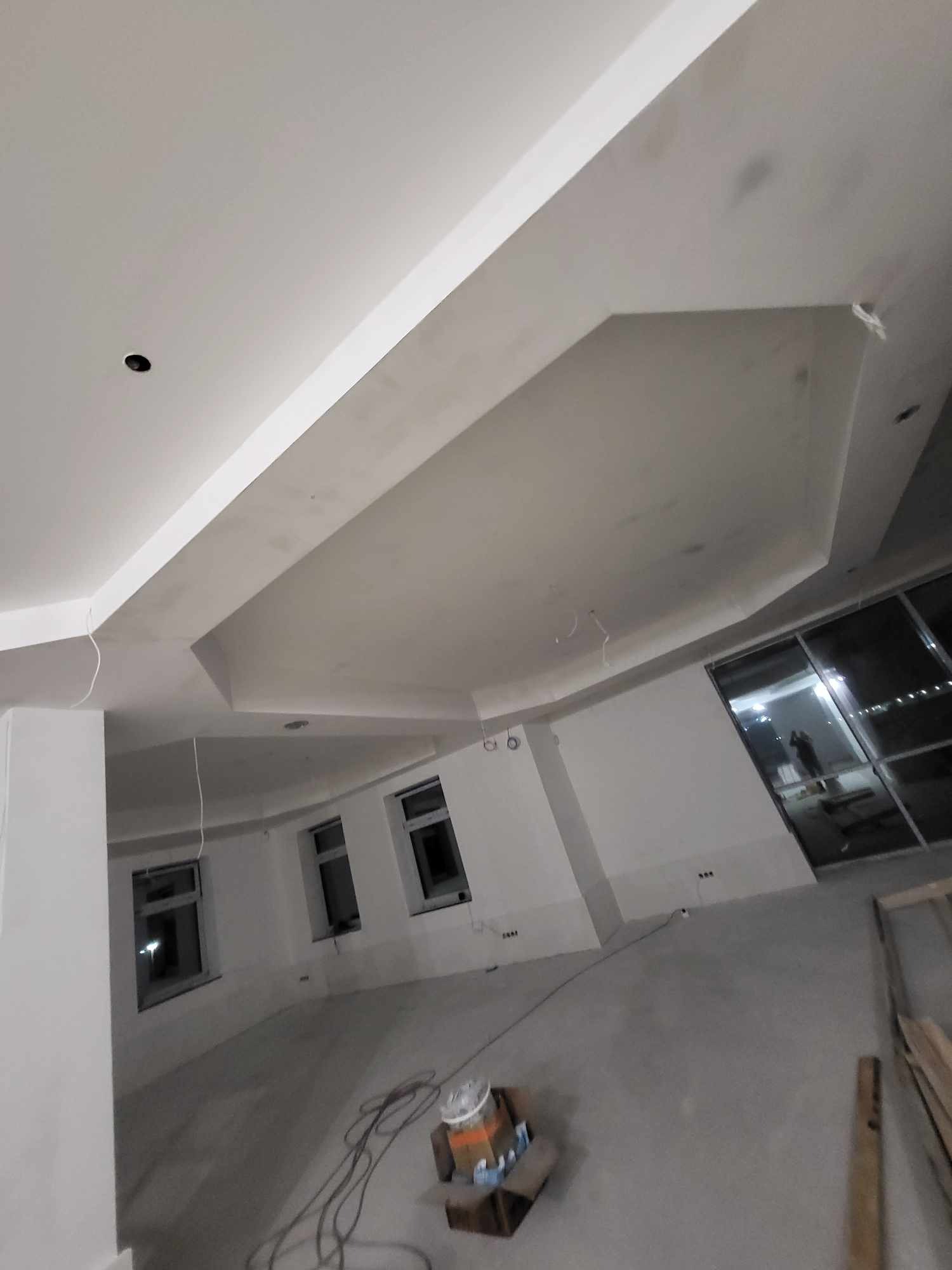 Wnętrze w stanie surowym: widoczny sufit z nietypową, kaskadową zabudową karton-gips, ściany przygotowane do malowania, okna z ciemnymi ramami, podłoga wylewka, kable zwisające z sufitu.