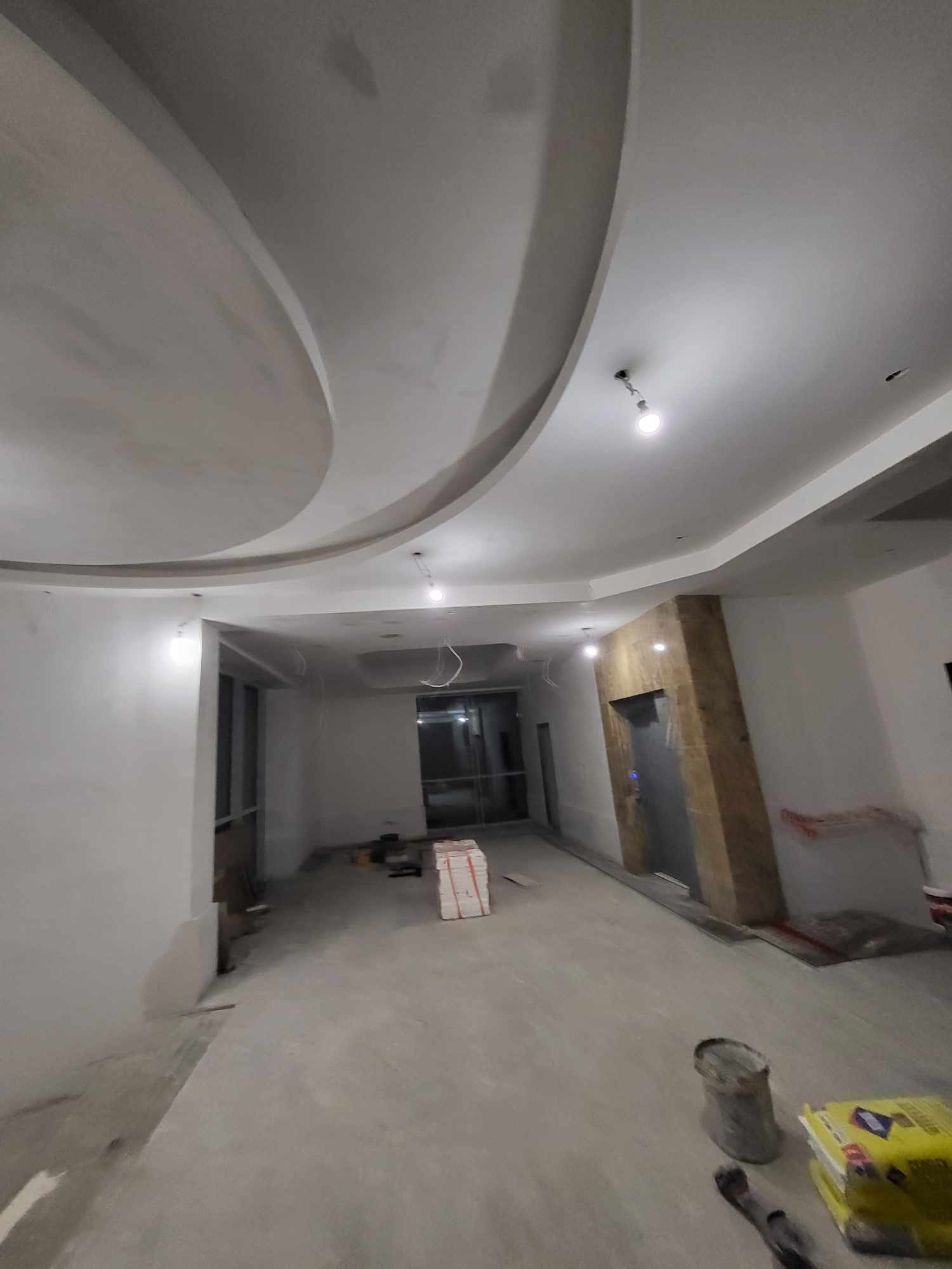 Wnętrze w trakcie prac wykończeniowych: surowy sufit z dekoracyjnymi elementami gipsowymi, ściany przygotowane do malowania, widoczne materiały budowlane i narzędzia na podłodze, fragment windy w tle.
