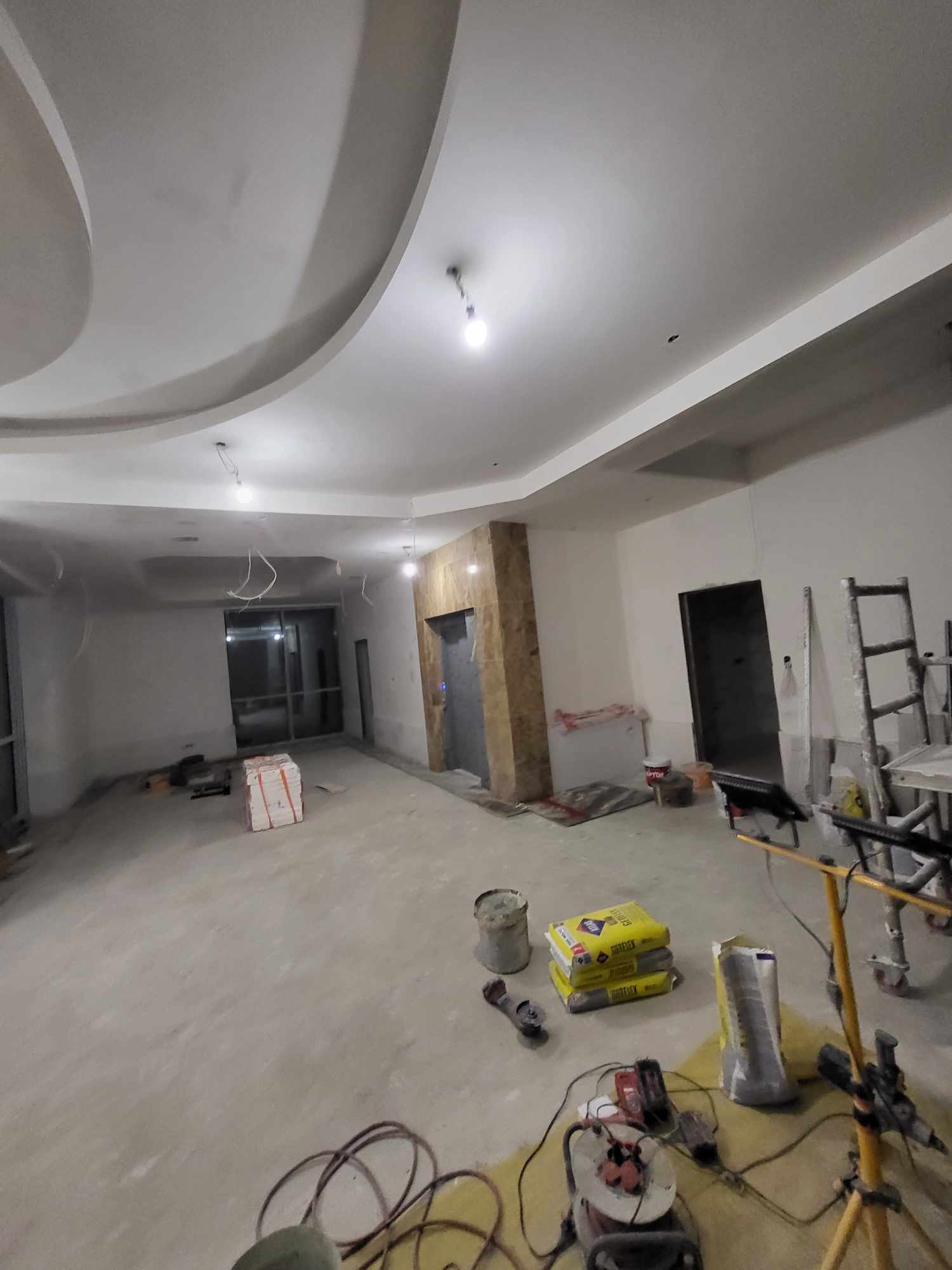 Wnętrze w trakcie prac wykończeniowych w Siemiatyczach: widoczny surowy sufit z dekoracyjnymi wgłębieniami, worki z zaprawą, narzędzia i drabina, sugerujące etap przygotowania do malowania...