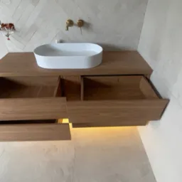 Drewniana szafka pod umywalkę z dwoma wysuniętymi szufladami i podświetleniem LED, umywalka nablatowa, bateria umywalkowa w kolorze złotym, widoczne kable instalacji elektrycznej.