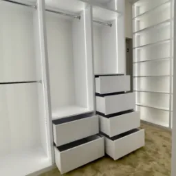 Nowoczesna, biała garderoba z oświetleniem LED, częściowo wysunięte szuflady, widok na brązowy dywan.