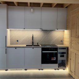 Vlad Tracenko - Jasna kuchnia w domku z bali: szare szafki z czarnymi uchwytami, blat imitujący marmur, czarna bateria i płyta indukcyjna, oświetlenie LED pod szafkami, widoczna drewniana konstrukcja ścian.