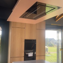 Vlad Tracenko - Nowoczesna kuchnia z jasnymi drewnianymi szafkami, różowym sufitem z wbudowanym okapem i wyspą kuchenną z widocznym wkładem na sztućce, widok na zewnątrz przez duże okno.