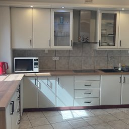 Vlad Tracenko - Jasna kuchnia z białymi szafkami, drewnianym blatem i szarymi płytkami, widok na okno z żaluzjami i czerwony czajnik.